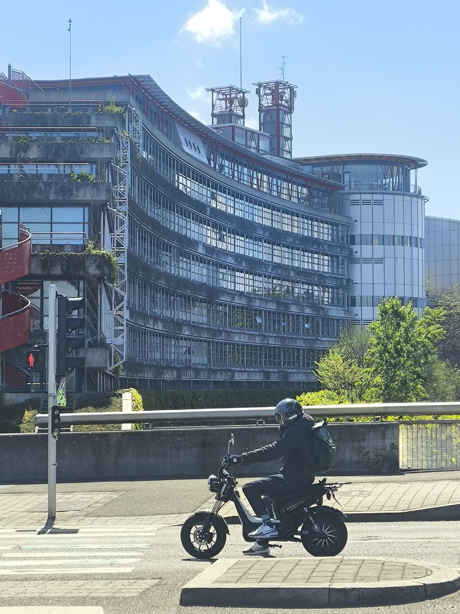 The image captures an urban street scene on a bright, sunny day, with a man riding an electric moped in the foreground. The man is wearing a dark helmet, a dark jacket, dark pants, and a dark backpack. His light-colored sneakers feature "AIR MAX" branding. The black moped has a prominent headlight and wheels, with "E-LUX" visible on its side panel. He is positioned on the street, near a pedestrian crossing.
In the background stands a distinctive, multi-story modern building with a curved glass and concrete facade. Its numerous windows reflect the sky, and several levels incorporate terraced balconies with some greenery. A red spiral staircase is visible on the left side of the building, which also features red structural elements and rooftop antenna towers. Adjacent to it on the right is a cylindrical building with silver paneling and round windows. Green trees and bushes grow around the base of the buildings and along a concrete barrier that spans the mid-ground.
A traffic light pole on the left displays a red 'don't walk' signal. The sky is clear blue with a few scattered white clouds, indicating fair weather and daytime. The setting is an urban environment in Strasbourg, France.