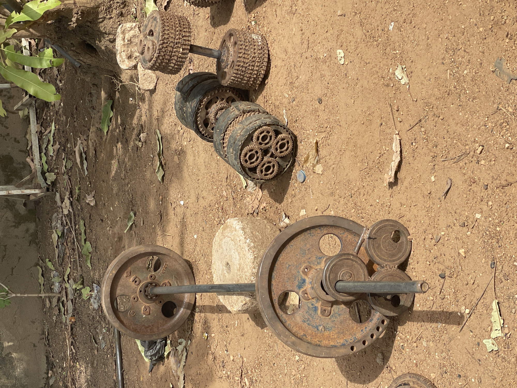 The image captures a collection of heavily rusted, makeshift weightlifting equipment resting on dusty, light brown ground under bright sunlight. The scene appears to be outdoors, with scattered small stones, dried leaves, and some green foliage visible along the left edge, possibly from a tree or shrub. The setting is consistent with an outdoor environment in Akwanga, Nigeria.
The weights are clearly repurposed and custom-made, showcasing a resourceful approach to gym equipment. In the upper-central part of the frame, a dumbbell is constructed from two large, heavily rusted, spiky gears or industrial wheels attached to a metal bar. Below this, a cylindrical weight lies on the ground, appearing to be made from stacked rubber rings or old tires, with an exposed end revealing five smaller, rusty gears arranged in a circular pattern.
In the lower half of the image, a barbell extends across the frame. It features two large, circular, heavily rusted weight plates on either end of a metal bar. One of these plates clearly displays the text "40LB," indicating its weight. A rough, light-colored stone or concrete block is positioned near the center of the barbell, possibly as a support or an additional weight. On the right side of the barbell, several smaller, rusty circular plates are stacked, adding to the weight assembly.
The pervasive rust on all metal components suggests prolonged exposure to the elements. Distinct shadows are cast by the equipment, indicating a clear, sunny daytime. There are no people visible, and no specific activity is taking place; it appears to be a static scene of stored or discarded weights.
