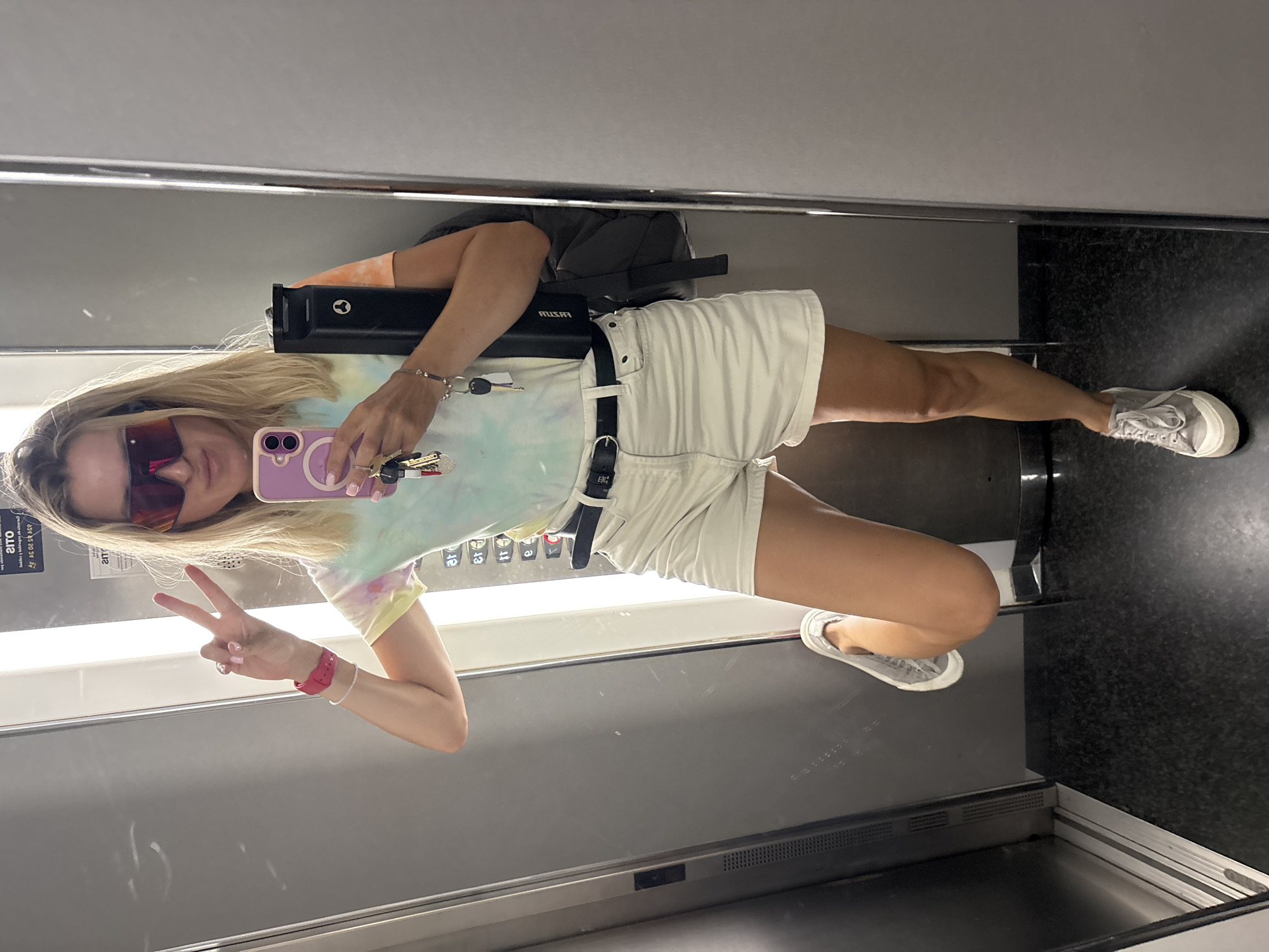 The image shows a young woman in an elevator in València, Spain. She is taking a selfie, holding up her phone to capture her reflection. She is wearing a tie-dye t-shirt, white shorts, a black belt, and grey sneakers. She has blonde hair and is wearing sunglasses. Her left hand is raised in a peace sign, and her right hand is holding her phone and a set of keys. A black bag is visible on her back. The elevator has a mirrored wall, reflecting the woman and the interior of the elevator. The lighting suggests it is daytime. There is text visible on a sticker inside the elevator, though it is mirrored and partially obscured. The woman appears to be in good spirits, with a relaxed posture and a playful gesture.