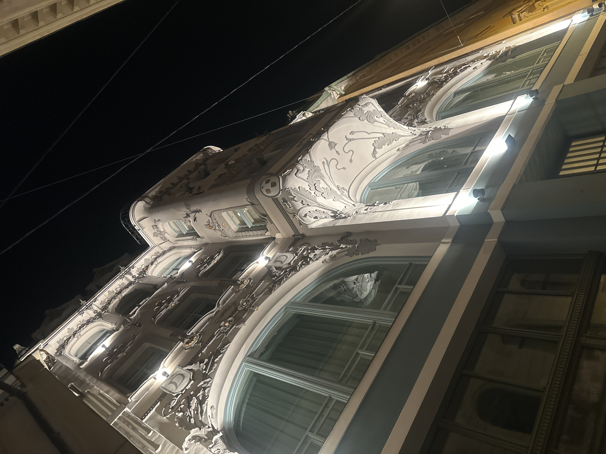 The image depicts the Art Nouveau architecture of a building in Riga, Latvia, at night. The perspective is from below, looking up at an angle. The building features ornate facade details, including floral and curvilinear elements characteristic of the Art Nouveau style. Large windows with rounded tops are prominent. Streetlights illuminate the facade, creating bright spots and casting shadows. The sky is dark, indicating it is nighttime. Electrical wires can be seen crossing the sky. The building's facade is likely made of light-colored stone or plaster.