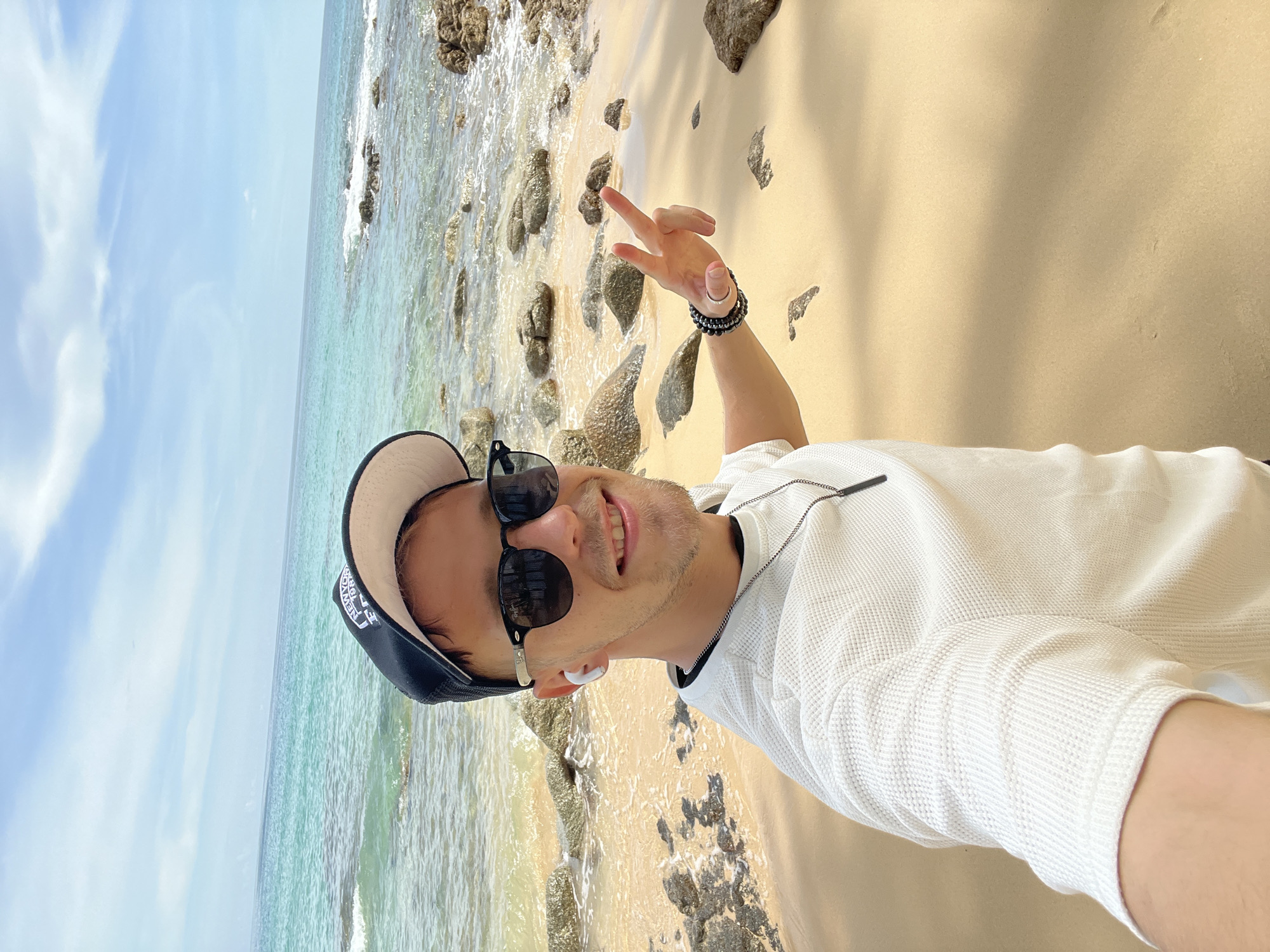 A man is taking a selfie on a beach in Choeng Thale, Thailand. He is wearing a white shirt, sunglasses, and a black baseball cap. He has on white earbuds and two bracelets on his left wrist. He is smiling and holding up two fingers in a peace sign. The background shows a sandy beach with rocks, and the ocean with turquoise water and a blue sky with clouds.