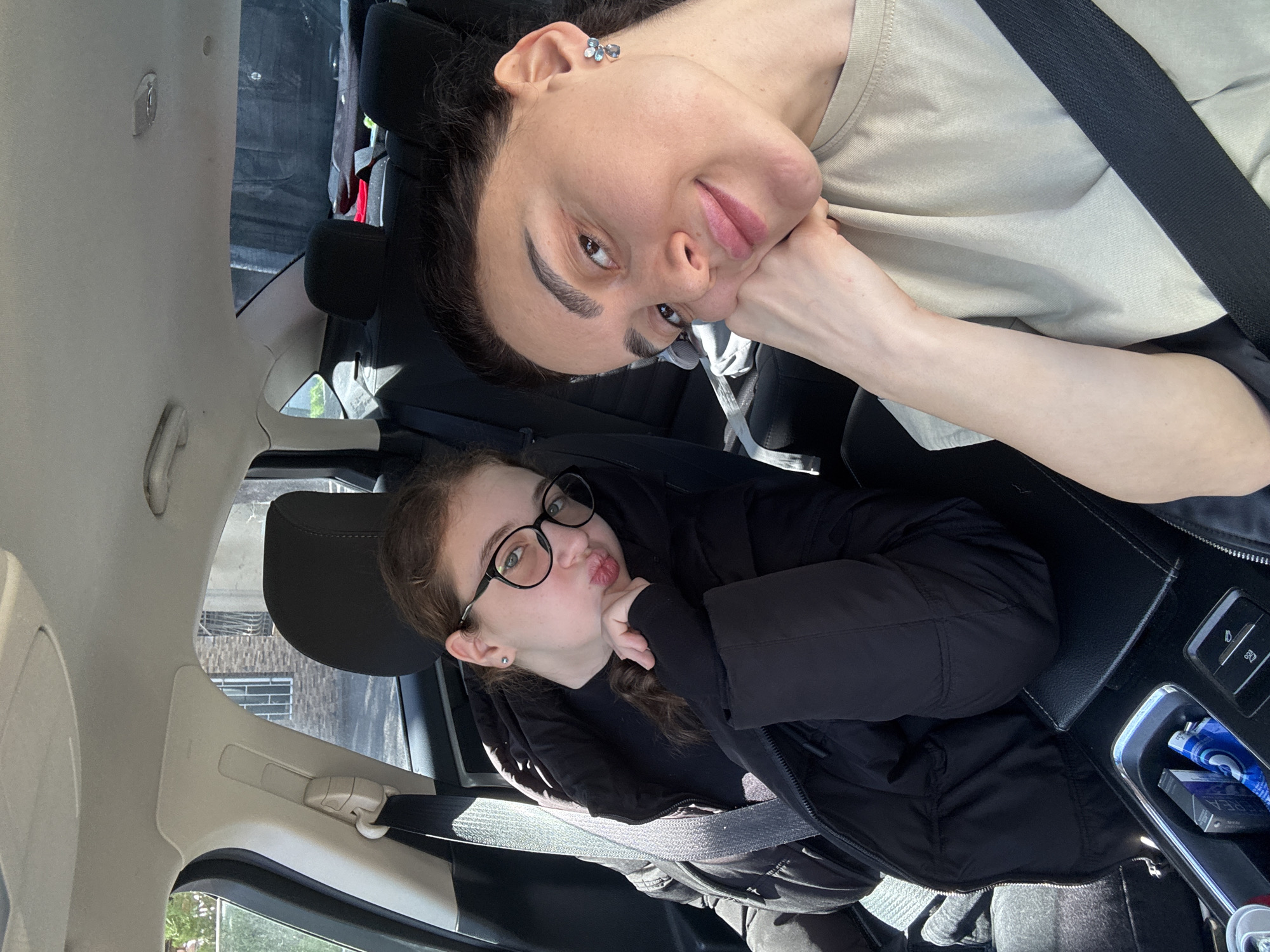Two females are inside a car. The older female is at the top of the image, wearing a seatbelt and a light beige shirt. She has dark hair pulled back, dark eyebrows, fair skin, and is resting her face on her hand. She is looking at the camera. The younger female is below her, also wearing a seatbelt and a black jacket. She has glasses and her hair in pigtails. She is making a kissing face with her lips puckered. The interior of the car is visible, including the dashboard and ceiling. Through the windows, a building can be seen, suggesting they are likely in Tbilisi, Georgia.