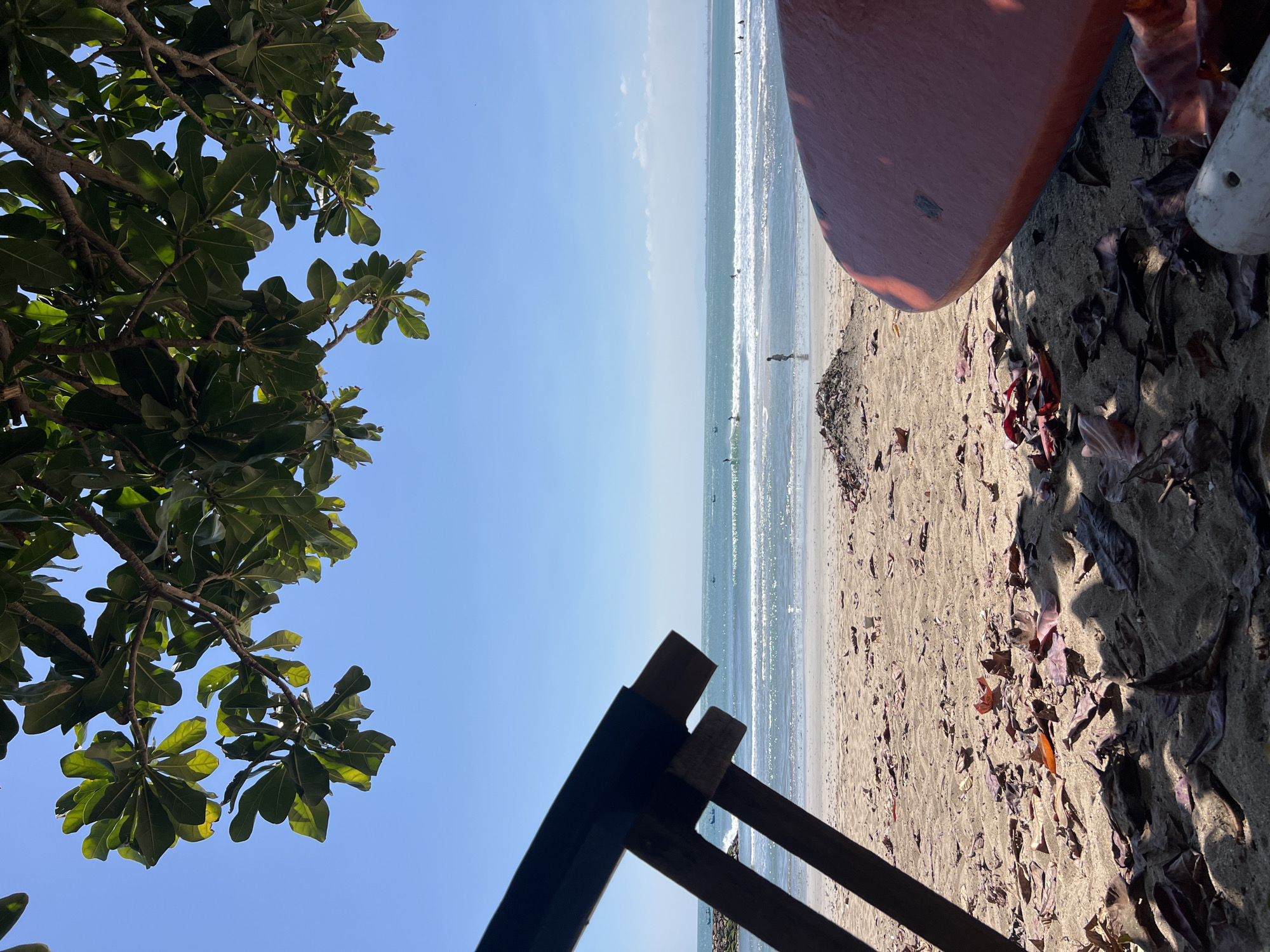 An eye-level shot captures a beach scene in Kuta, Indonesia. The foreground is dominated by sand littered with fallen leaves, with a section of an orange surfboard angled in the lower right corner and the dark frame of a beach chair on the left. Beyond the sand, gentle waves roll onto the shore under a clear blue sky. Several surfers are visible in the ocean, and a lone figure walks along the water's edge. Lush green foliage from an overhanging tree frames the top of the image.