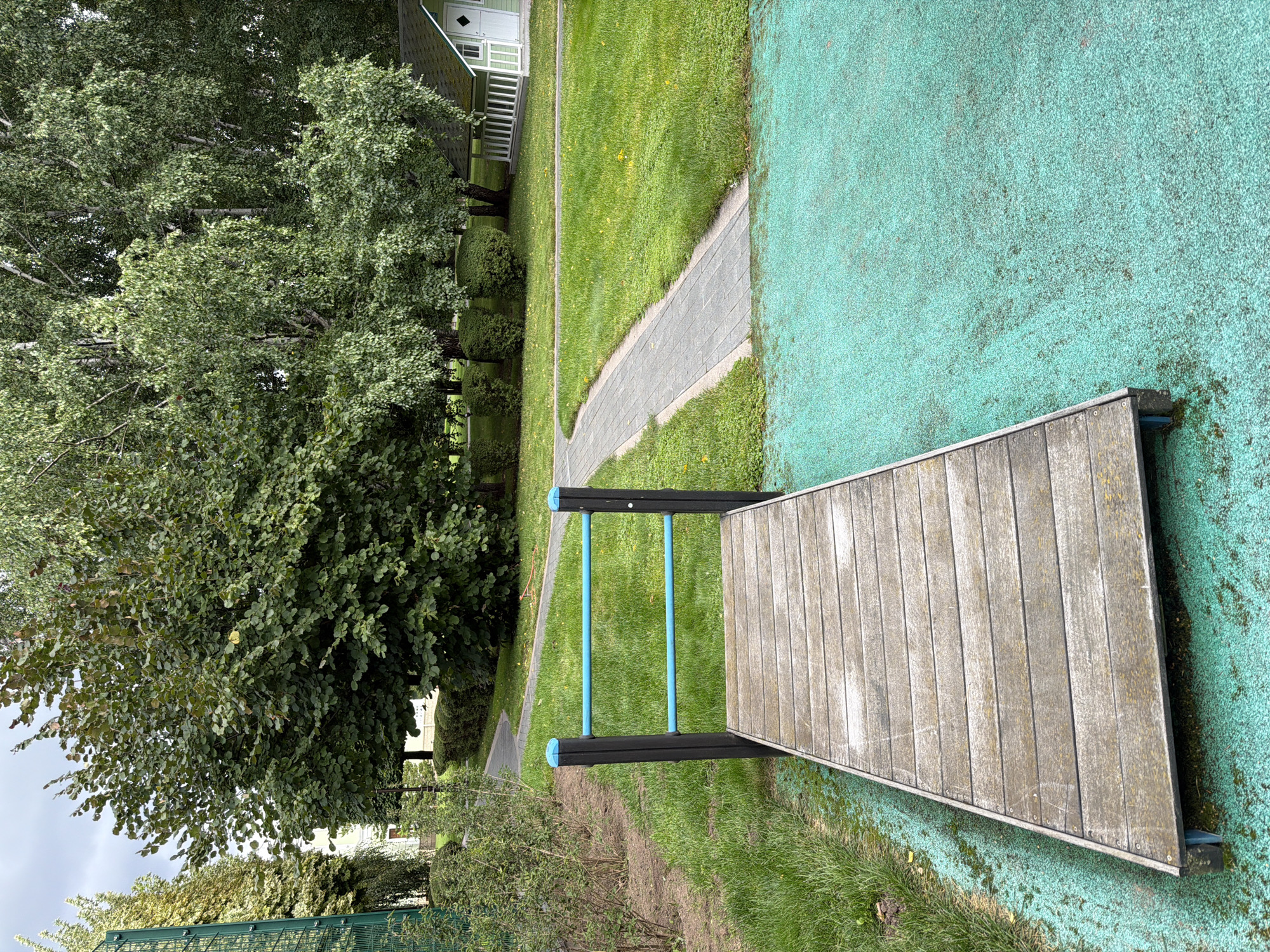 The image depicts an outdoor scene in Zhukovka, Russia, featuring a wooden ramp with blue railings, positioned on a textured turquoise surface. The ramp leads towards a paved pathway that curves through a well-maintained grassy area. In the background, a small, white-trimmed green house with a dark roof is visible amidst lush trees. The environment is predominantly green, with a mix of mature trees and manicured bushes, under an overcast sky.