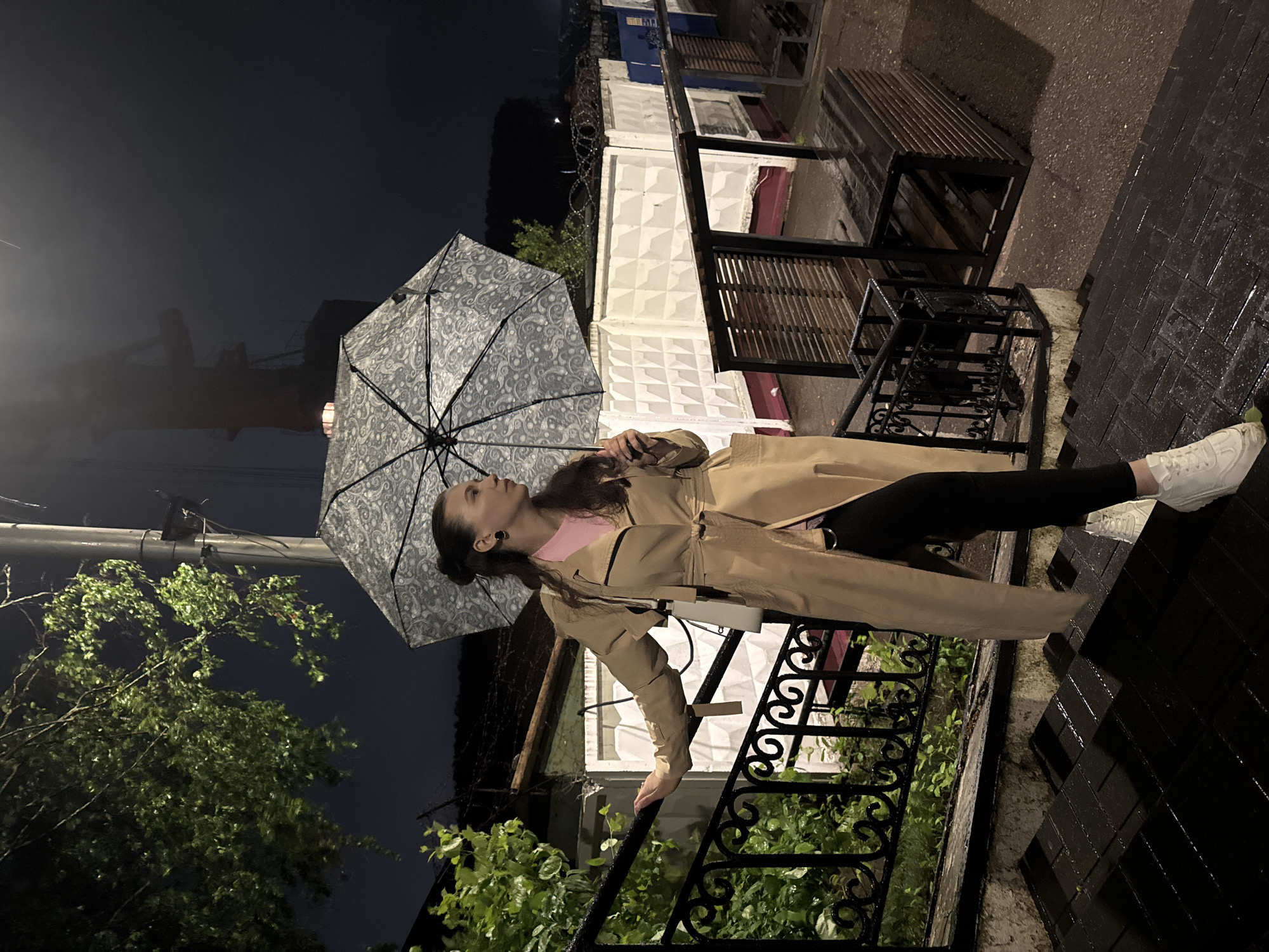 The image captures a female individual standing on wet, dark-colored outdoor steps at night in Chaikovsky, Russia. The subject has dark hair styled in a partial bun and wears a light tan trench coat, a pink top, black leggings, and white sneakers. Her right hand rests on a black ornate metal railing, and her left hand holds an open umbrella featuring a grey and white paisley-like pattern. She is looking towards the right side of the frame.
The environment indicates recent precipitation, with visible moisture on the steps, railing, and surrounding surfaces. To the left, green foliage from trees and bushes is present. In the midground, a light-colored building or wall is partially visible. Further right, a white concrete fence with a repetitive geometric pattern is topped with barbed wire, behind which a dark blue corrugated structure and a wooden outdoor bench or seating area are located. The far background reveals a dark night sky with a silhouetted industrial structure, possibly a crane, showing faint red-orange illumination near its base, and a utility pole. Ambient artificial lighting is present, reflecting off the wet surfaces.
