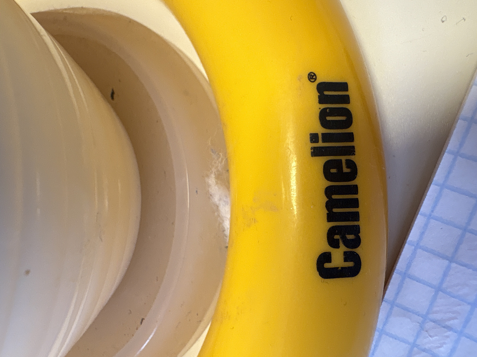 The image shows a close-up view of a bright yellow object with black text that reads "Camelion" followed by a registered trademark symbol. The yellow object appears to be a curved, tubular structure. The background is out of focus, revealing a light-colored, curved surface and a piece of graph paper in the lower left corner. The scene is indoors, and there are no people or animals visible. There is no discernible connection to the city of Georgiyevsk, Russia, or any specific location within it.