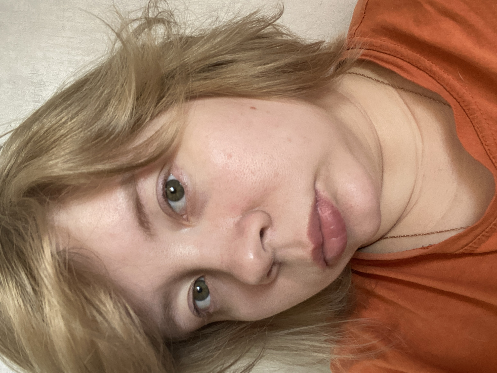 A close-up, angled portrait of a light-skinned person with light brown, wavy hair. The person is wearing an orange t-shirt and a delicate gold necklace. The person's eyes are green. The background appears to be a light-colored surface, possibly a bed or couch. The location cannot be determined with the provided image.