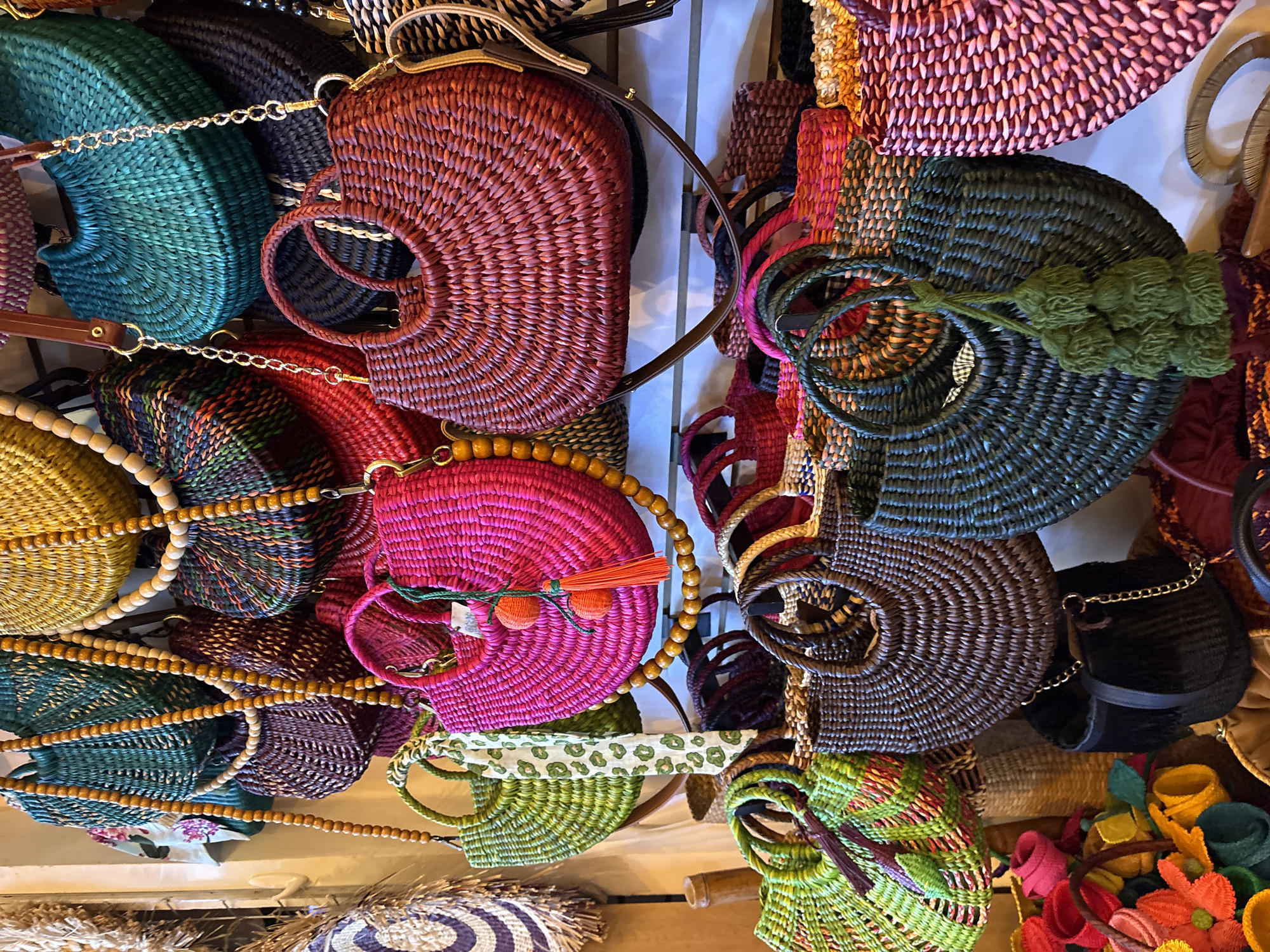 This image depicts a vibrant display of woven straw and raffia handbags at a market stall, likely in Miraflores, Peru. The scene is densely packed with a variety of bags in an array of colors and sizes, hung from a white wall or display structure.
Dominating the upper portion of the frame are several large bags. A bright fuchsia bag with a beaded strap and orange tassel is prominently featured. To its left, a lime green bag with a leopard-print scarf tied to its handle is visible. Above these, a large, dark teal bag with a chain strap hangs, and to its left, a deep yellow woven bag with a beaded strap. Other bags in shades of green, brown, and red are also visible in the upper and middle sections.
The lower part of the image reveals a dense collection of smaller handbags and baskets. A large dark brown bag with a rounded shape and braided handles is in the center. To its right, a dark teal bag with a green tassel hangs. Several smaller bags in pink, green, and brown tones are stacked or arranged in front of the larger bags. On the far left, a basket filled with colorful woven flowers adds a decorative touch.
The overall impression is one of abundance and artisanal craftsmanship, typical of a local market. The lighting is bright, highlighting the textures and colors of the woven materials. No people are visible in the frame.