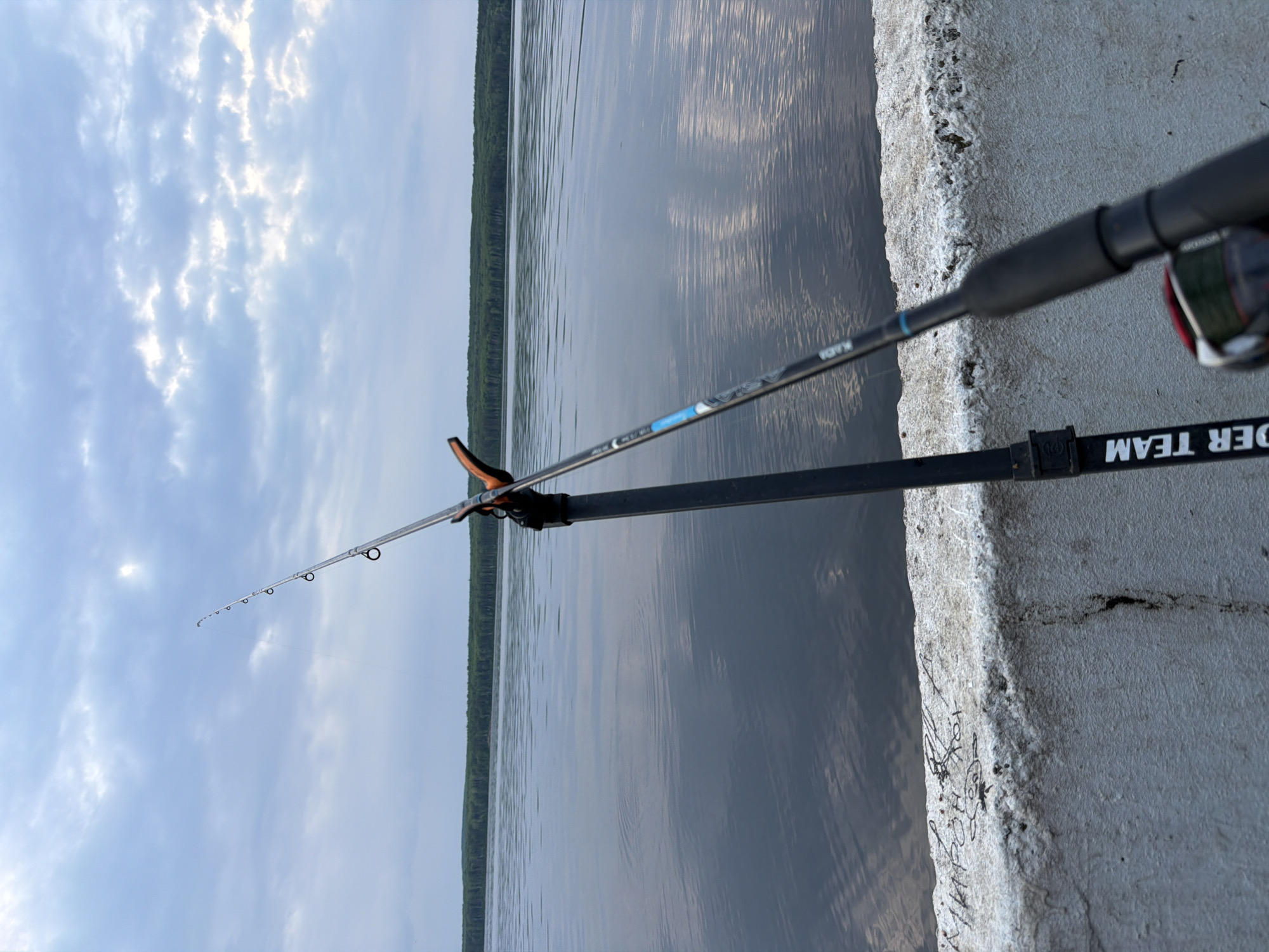 The image captures a peaceful fishing scene by a body of water in Chaikovsky, Russia. A fishing rod with a reel is set up on a black stand, positioned over the water. The rod displays text such as "KAIJU" and "SOFTY", while the stand features "RIDER TEAM".
The fishing gear rests on a grey, weathered concrete structure, likely a pier or embankment, which shows signs of age and includes some indistinct graffiti, such as scribbled drawings and text. The water's surface is calm with gentle ripples, reflecting the sky. Across the water, a dense, dark green forest lines the distant shore. The sky is partly cloudy, with soft, diffused light, suggesting either early morning, late afternoon, or an overcast day. The scene evokes a sense of tranquility and the patient activity of fishing.