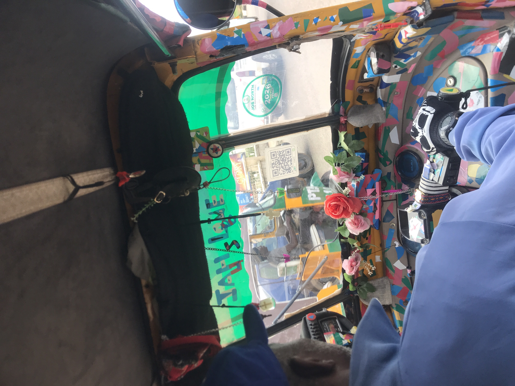 The image shows the interior of a brightly decorated auto-rickshaw, or "keke," in Jos, Nigeria. The driver, wearing a blue shirt and a blue baseball cap, is visible from the back left, facing forward. The auto-rickshaw's interior is covered in colorful, abstract patterns and numerous stickers. A green fabric canopy is stretched across the top, with the word "JAH LOVE" written on it in large letters. Artificial flowers, including red and pink roses, are attached to the dashboard. In the background, other vehicles are visible, including a blue truck and a white van with a sticker that reads "JOS NORTH LOCAL GOVERNMENT COUNCIL 2026 TRICYCLE COUNCIL." The overall scene depicts a busy street environment in Jos, Nigeria, characterized by the vibrant decoration of the auto-rickshaw and the presence of other traffic.