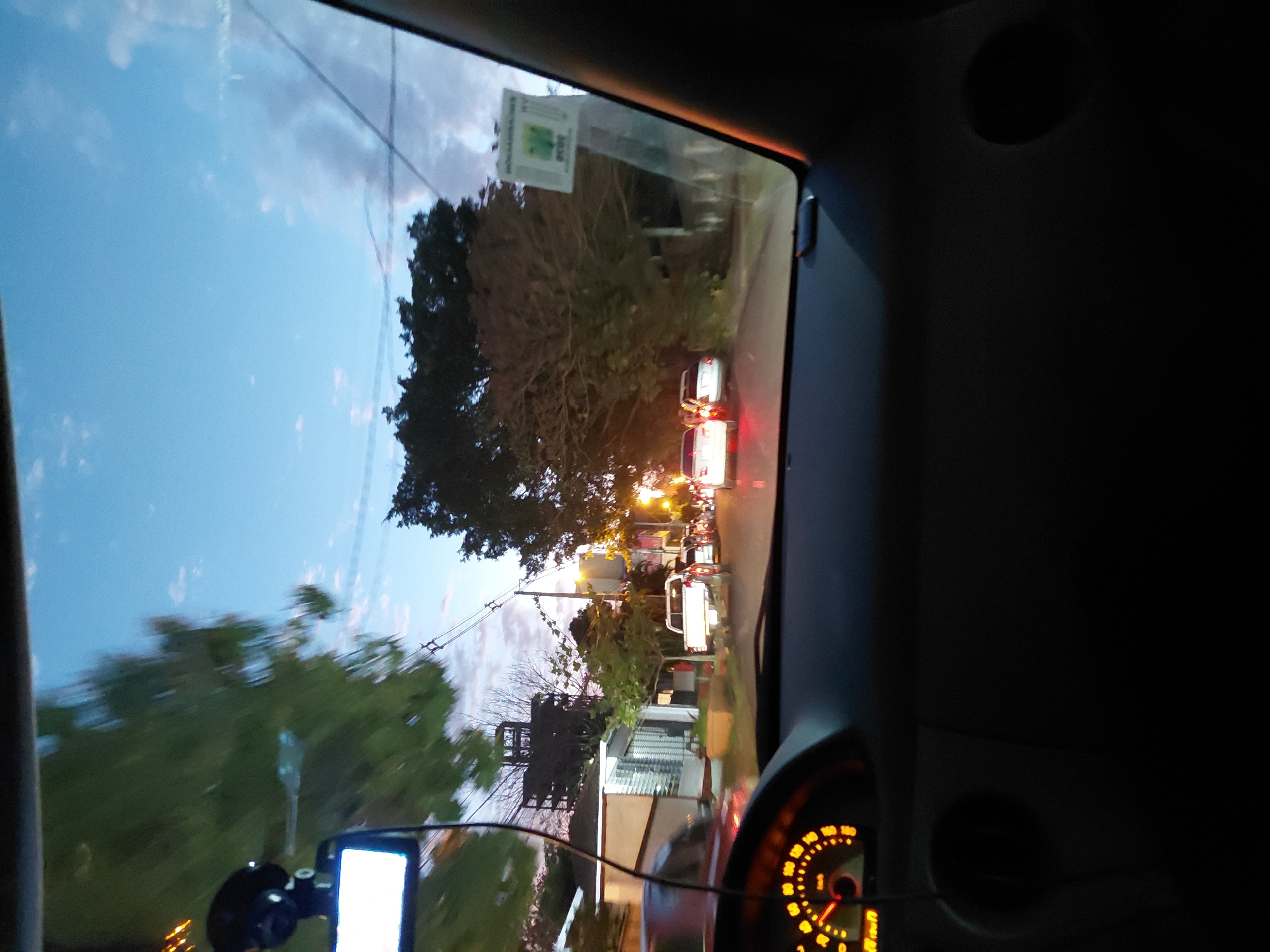 The image is taken from inside a vehicle, looking forward through the windshield at dusk. The sky is a gradient of blue and purple, with scattered clouds. The road ahead is lined with trees and buildings. Several cars are visible on the road, some with their headlights on. The dashboard of the vehicle is partially visible in the foreground, with an illuminated speedometer. The scene appears to be a residential or semi-urban area. This photograph was captured at the intersection of General Máximo Santos and Monseñor Wiest streets in Encarnación, Paraguay.