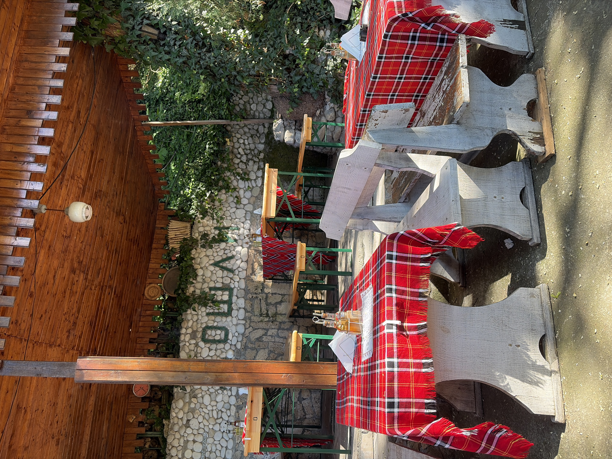 The image shows an outdoor seating area, likely at a restaurant, located in the city of Unknown, Unknown. There are several wooden tables covered with red plaid tablecloths. The tables are accompanied by green metal chairs. The seating area is set against a backdrop of a stone wall with some greenery. A wooden structure is visible on the left side of the image. A clock is mounted on the wall behind.
