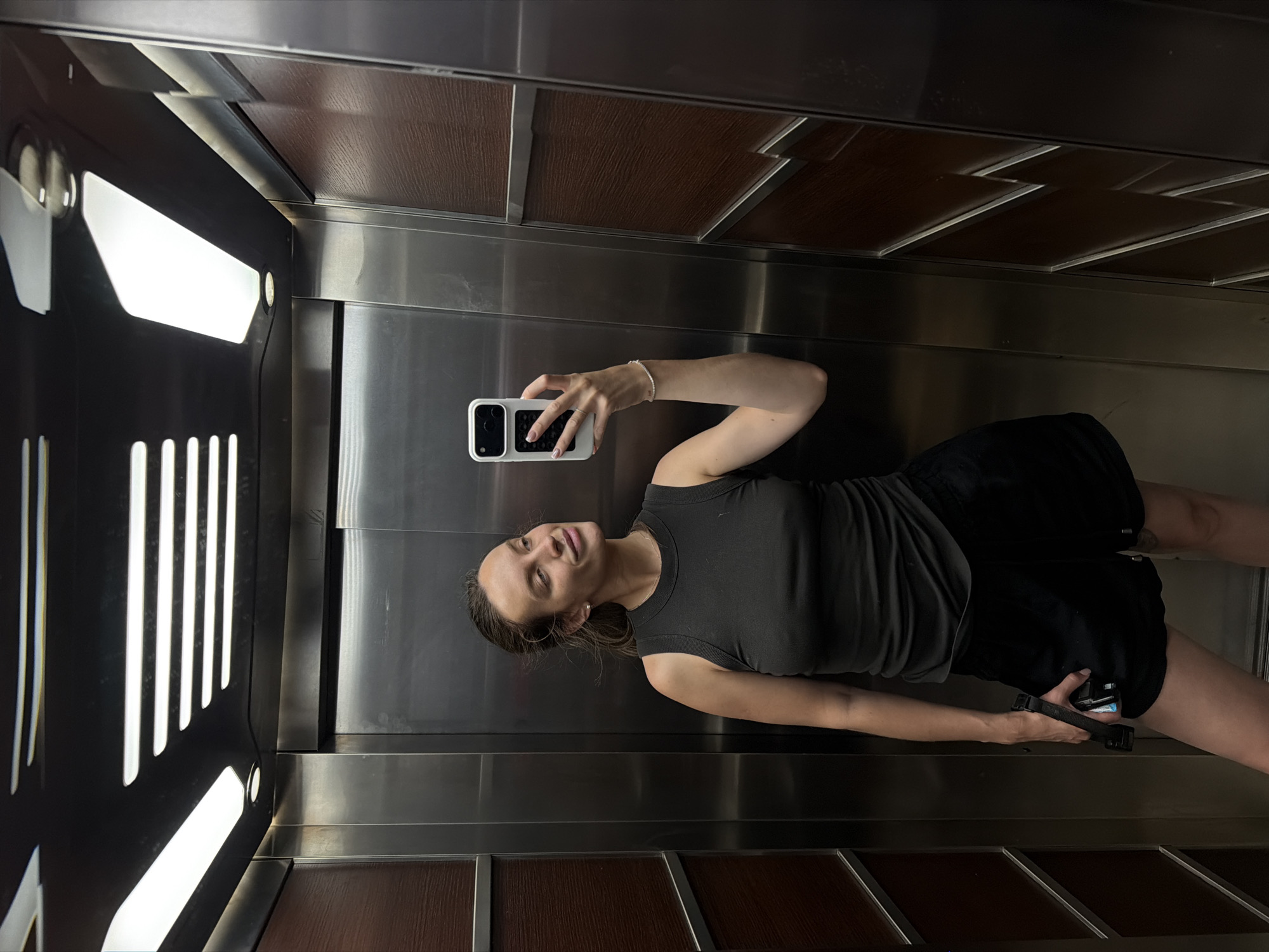 A young woman is taking a mirror selfie in what appears to be an elevator. She is wearing a dark grey tank top and black shorts. She is holding a white smartphone with a black case in her right hand, and her left arm is resting on her hip. The elevator walls are made of polished stainless steel and dark wood panels. There are bright light fixtures on the left side of the elevator. The woman is smiling and looking at her reflection. The overall mood of the image is casual and relaxed. There is no visible text in the image. The location is Budva, Montenegro.