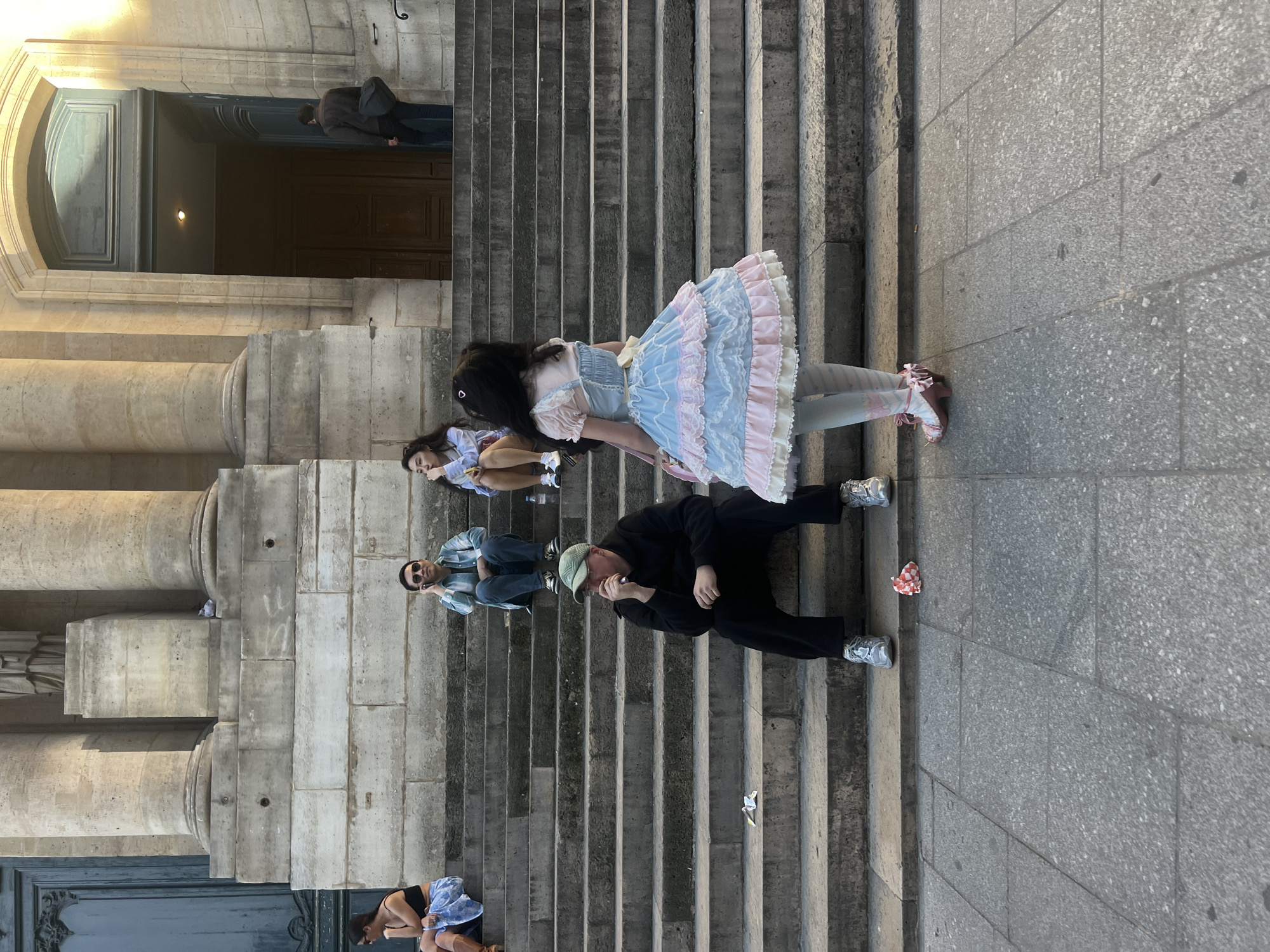 This image, captured in Paris, France, depicts a daytime outdoor scene on the grand stone steps of a large, classical-style building. The photo is oriented vertically, showing individuals casually sitting and resting on the wide staircase and adjacent paved areas.
In the mid-ground, a person wearing an elaborate light blue and pink ruffled Lolita-style dress, white blouse, patterned tights, and pink ballet-style shoes stands facing away from the viewer. Below them, another individual with dark hair sits on a step. To the right, two men are seated on the steps: one in a black long-sleeved shirt, dark trousers, silver sneakers, and a light green baseball cap, appearing to eat or drink; the other, behind him, in a striped blue and white shirt, dark trousers, and sunglasses, looking relaxed.
Closer to the foreground, a person in a dark top and light blue patterned fabric is seated on the lowest visible step. In the background, within a large arched stone doorway, another person in a dark jacket and shoulder bag is partially visible. The building's architecture features imposing stone columns and large, dark wooden doors, characteristic of historic Parisian public structures. Small pieces of litter, including a red and white checkered wrapper, are scattered on the steps.
The scene conveys a relaxed, casual atmosphere, suggesting people taking a break or enjoying the public space. The lighting indicates daytime, possibly late afternoon. No discernible text is visible.
