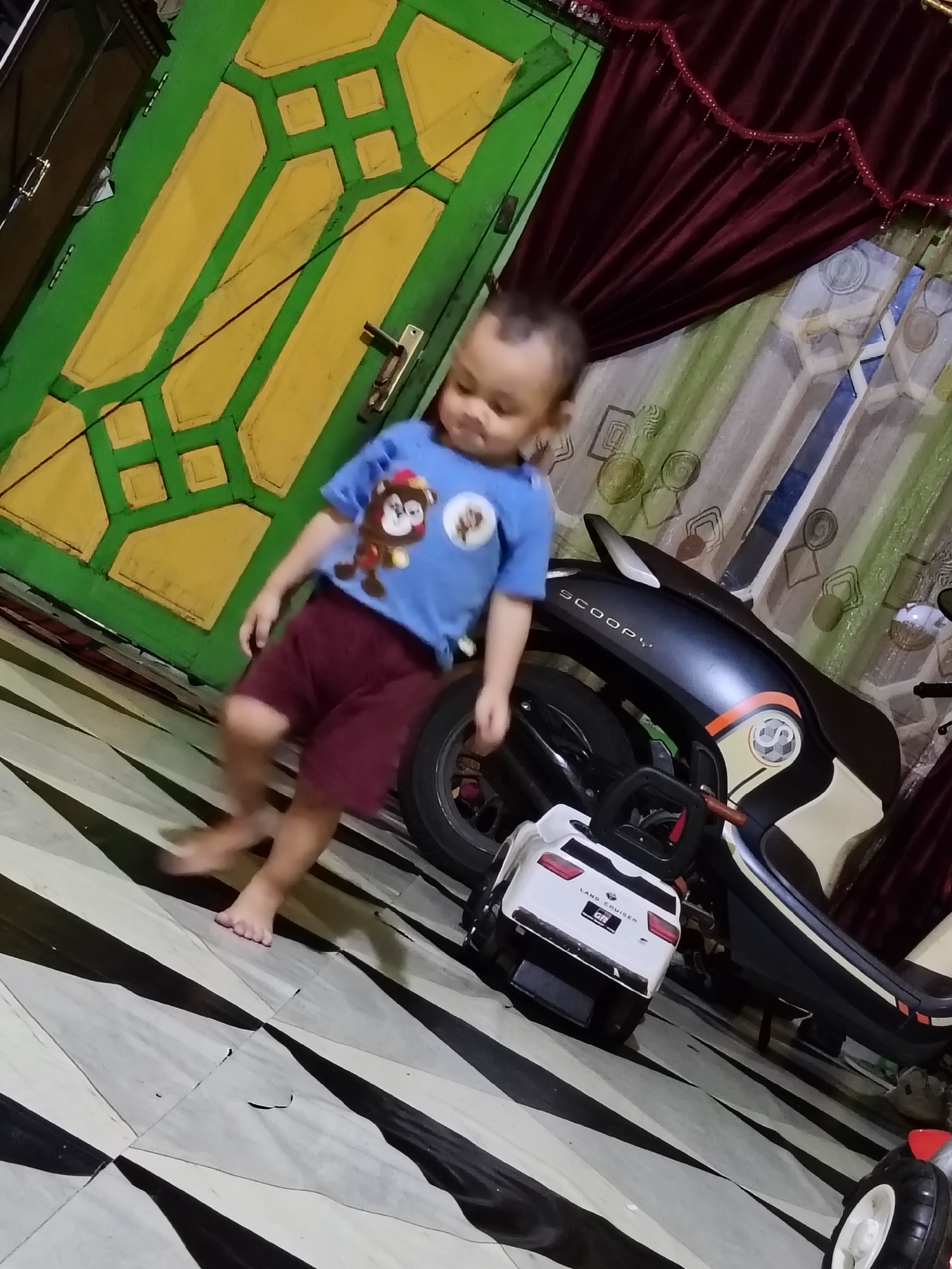 In a domestic interior in Tanah Grogot, Indonesia, a young child wearing a blue t-shirt with a monkey graphic and maroon shorts is captured mid-stride, its bare feet blurred due to motion. The child is near a white toy car, which has the text "LAND CRUISER GR" on its rear. Behind the toy car and child is a black and beige scooter with the word "SCOOPY" visible on its side. The environment includes a green and yellow door with decorative panels, dark red curtains, and a patterned floor with black and white diagonal stripes. The lighting suggests an indoor setting, possibly during the day with artificial light sources contributing to the illumination.