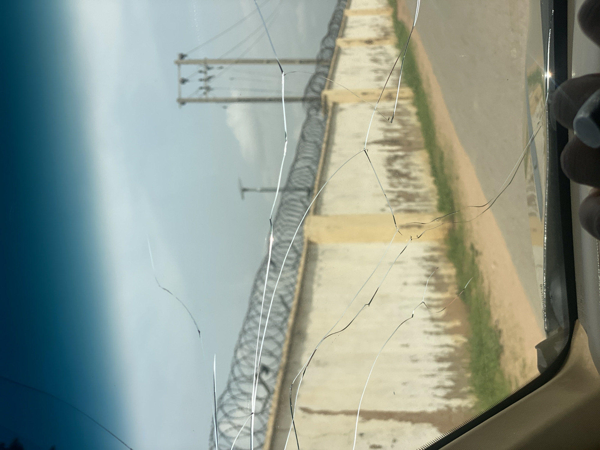 An exterior shot captures a cracked windshield view of a road leading towards a cream-colored wall topped with barbed wire. The wall, which appears to have some discoloration and texture, runs along the right side of the road, bordered by patches of green grass and dirt. In the background, a utility pole with crisscrossing wires is visible against a cloudy sky. The scene is observed from inside a vehicle, with the edge of the dashboard and a portion of a person's hand wearing a ring visible in the foreground. The image provides no specific details to indicate the location within Howayi, Nigeria.