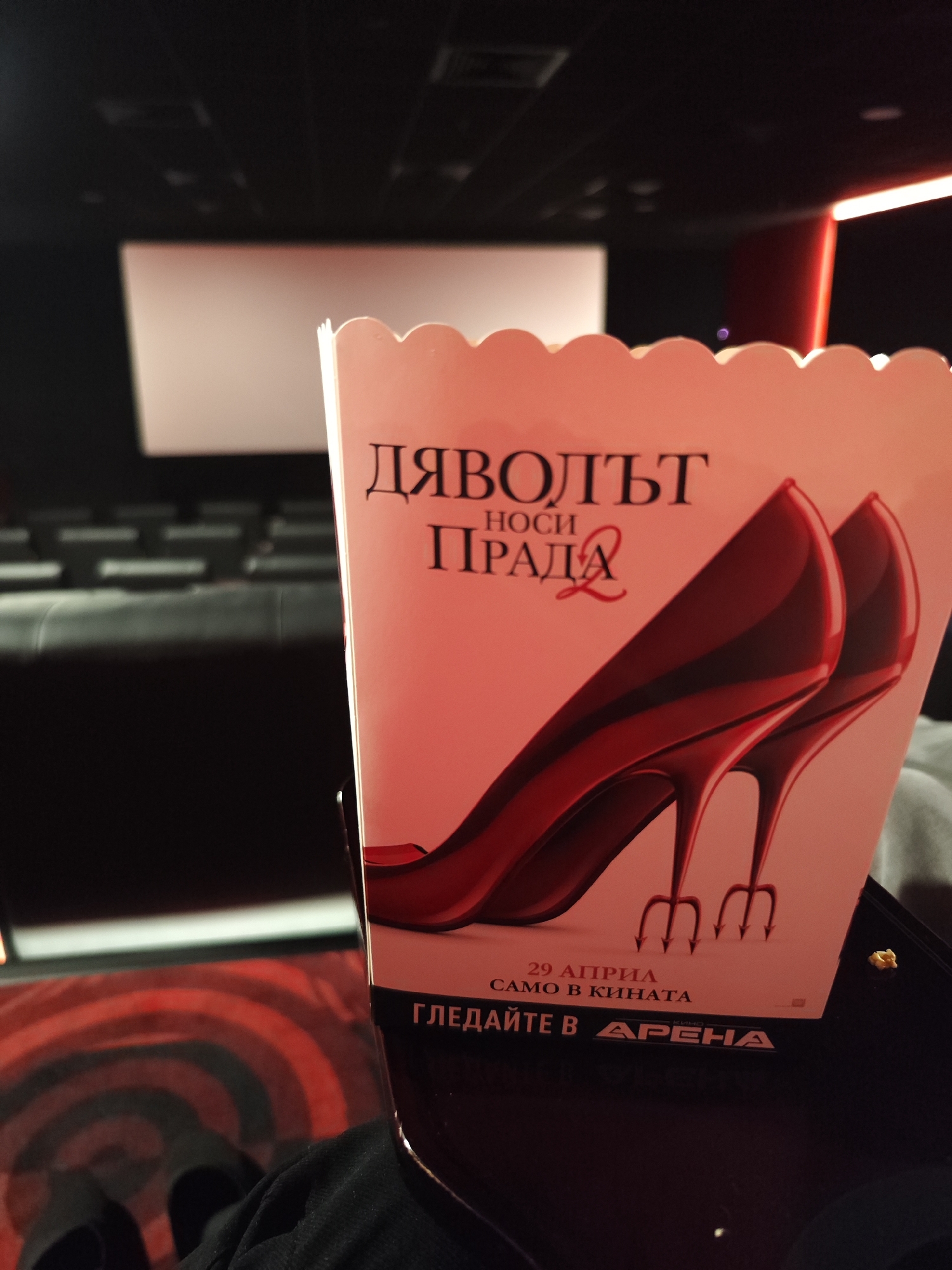 The image depicts a popcorn container for the movie "The Devil Wears Prada 2" in a movie theater. The container shows two red high-heeled shoes with the movie title in Cyrillic above. Below the shoes is the release date in Cyrillic "29 April" and the phrase "Само в кинота" (Only in cinemas). At the bottom is "Гледайте в Арена" (Watch in Arena). The background shows a cinema screen, empty seating, and red ambient lighting. Based on the movie advertisement and the location named "Arena", it is likely that the picture was taken in a cinema located in Sofia, Bulgaria.