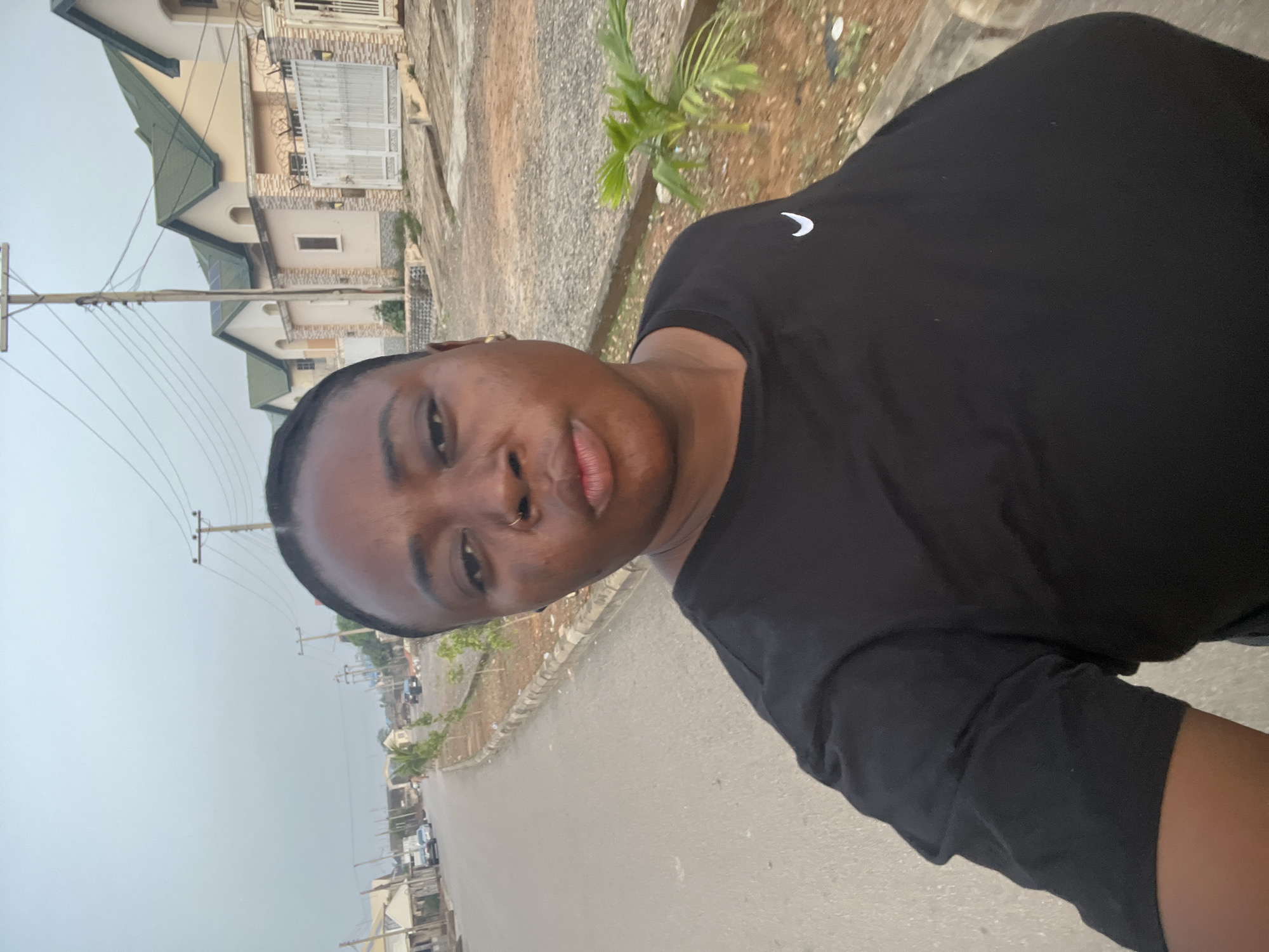 The image depicts a person with dark hair and a nose ring wearing a black t-shirt. The person appears to be standing on a paved street with houses and utility poles visible in the background. The sky is overcast. Based on the architecture and general environment, the location is likely a residential area in a city, possibly Abuja, Nigeria. There are cars parked along the side of the road.