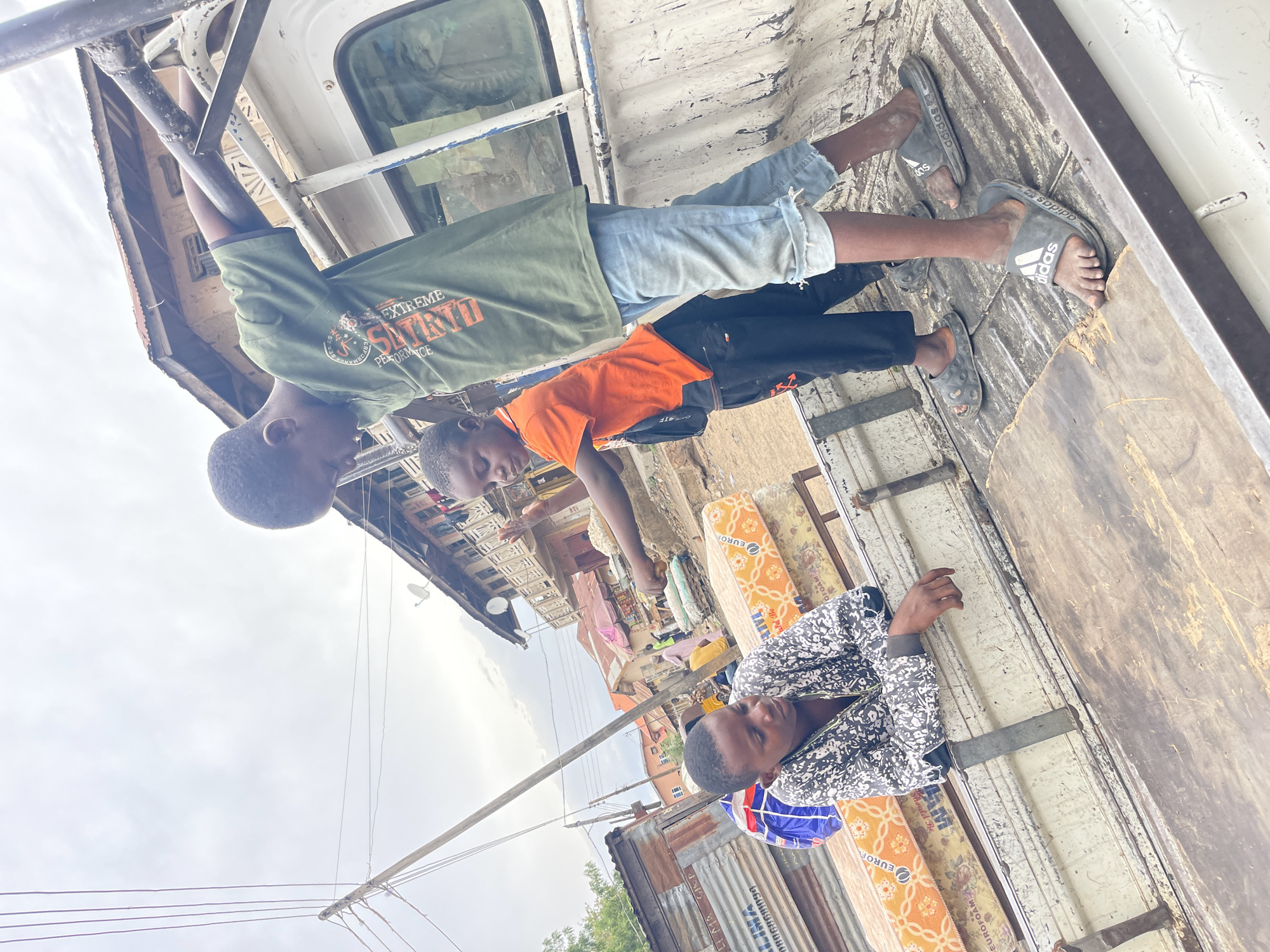 The image shows three individuals on what appears to be the back of a pickup truck, laden with foam mattresses. Two younger individuals, possibly children or adolescents, are positioned at the front of the truck bed. One is wearing an olive green shirt with the words "EXTREME PERFORMANCE SITINO" on it, and light blue denim shorts, while the other is wearing an orange shirt and dark pants, with Adidas slide sandals. They are holding onto the metal framework of the truck. A third, possibly older, individual is at the back wearing a white and black patterned jacket, lying beside some foam mattresses that have "EUROFOAM" printed on them. The truck bed appears to be made of wood, showing signs of wear.
In the background, there is a dense urban environment with buildings constructed from corrugated metal. Clothes hang on a line to dry, suggesting a residential area. Overhead, power lines stretch across the frame. The image likely comes from the city of Zaria in Nigeria, due to the printing on corrugated metal "LI MA SHOP 08068681094" a phone number that matches Nigeria country code +234