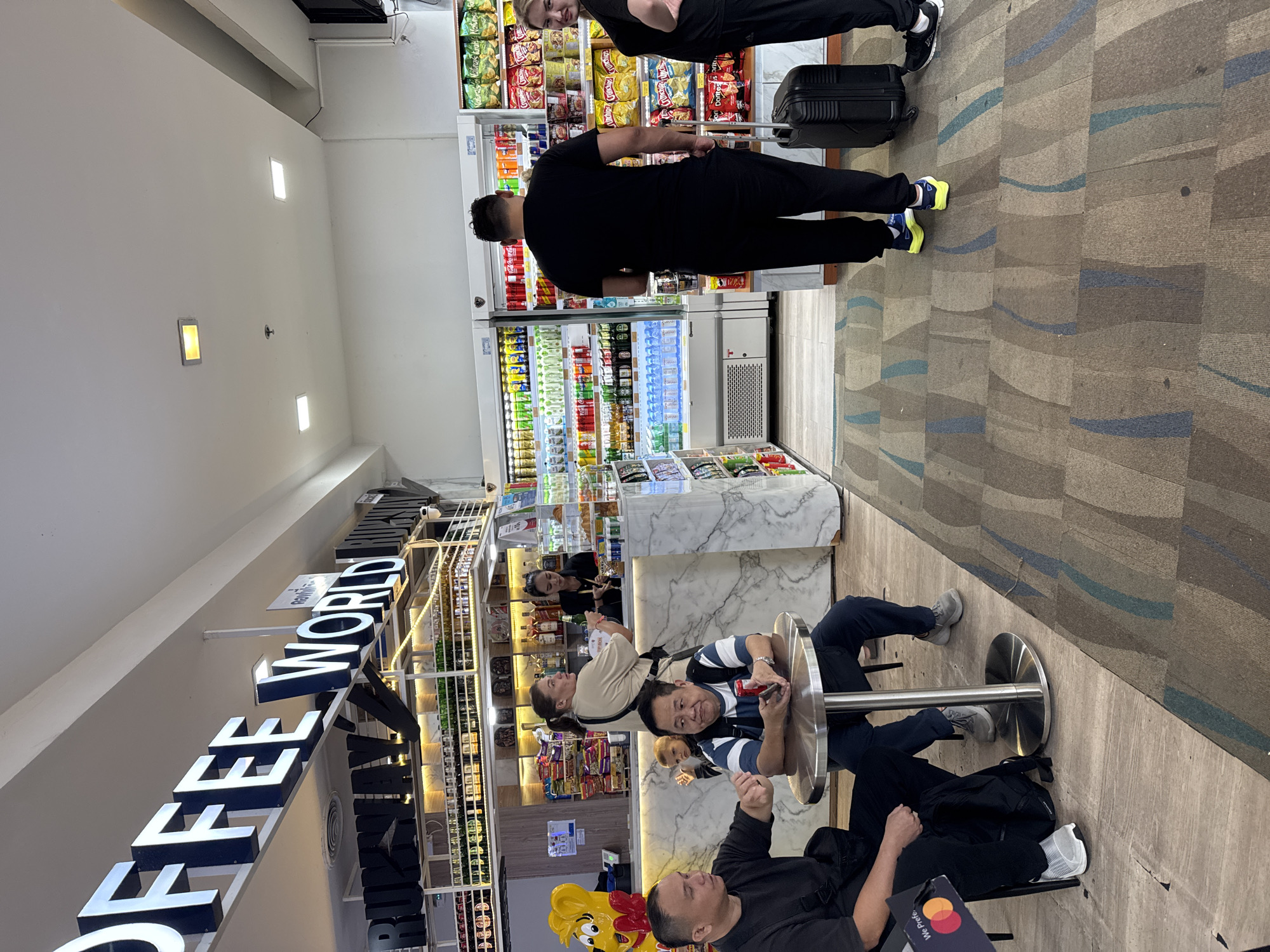 The image captures an interior view of a cafe and convenience store combination, identified by the overhead signage as "COFFEE WORLD RUNWAY," located within Phuket International Airport in Mai Khao, Thailand.
In the foreground to the left, two adult males are seated at a small, round metallic table on a light wood-patterned floor. The man on the left wears a black t-shirt and has a black shoulder bag, gesturing with his right hand towards the other man. The man on the right, wearing a striped polo shirt and a dark backpack, holds a red object, possibly a drink bottle or phone, and wears a watch on his left wrist.
Behind the seated men and to the left, a marble-patterned service counter is visible. A woman in a light-colored top stands in front of the counter, holding a green bottle and looking towards the right. Behind the counter, another woman in a dark uniform is present. A young child is partially visible between the seated men and the counter, looking right. On the far left of the counter, a large yellow cartoon chicken-like figure with a red bow tie stands. Above the counter are shelves displaying various bottles and packaged snacks.
To the right, a convenience store section features multiple glass-front refrigerators filled with a wide assortment of bottled and canned beverages. Next to these, open shelves are stocked with numerous packaged snack foods, including various brands of chips and instant noodles. A man in a black t-shirt and black pants stands with his back largely to the viewer, facing the refrigerated shelves. A black roller suitcase rests beside his left leg. To his right, partially visible, is another person in a dark top.
The flooring transitions from the wood-patterned tile in the cafe area to a patterned carpet with abstract brown, blue, and gray shapes in the standing area. The ceiling is white with recessed lighting fixtures. The overall scene is brightly lit and appears to be a busy commercial space.