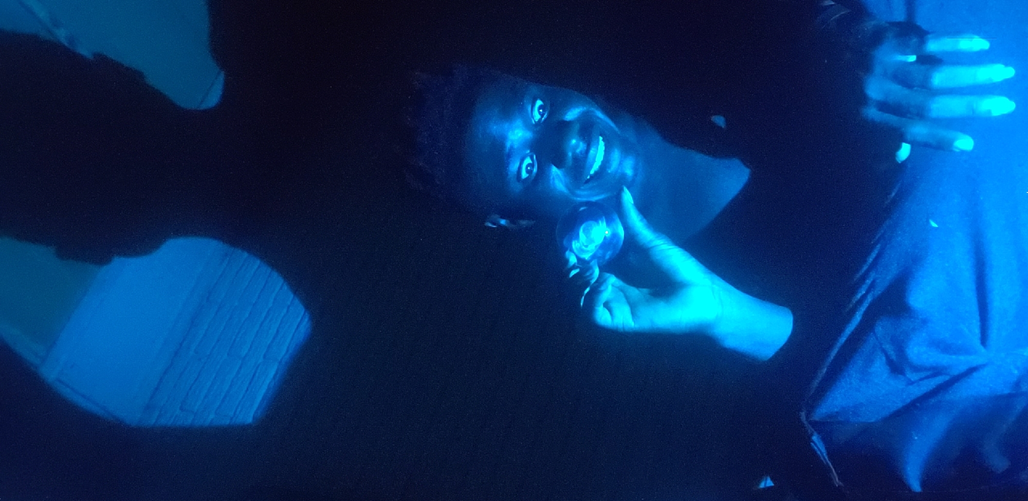 A person with dark skin and short hair is holding a small, glowing, spherical object in their right hand, looking directly at the viewer and smiling. The person is wearing a dark-colored shirt. Their left hand is raised near their face. The scene is illuminated with a strong blue light, which casts strong shadows. A rectangular object with a ribbed surface is visible in the lower left corner. The location is not apparent based on the image.