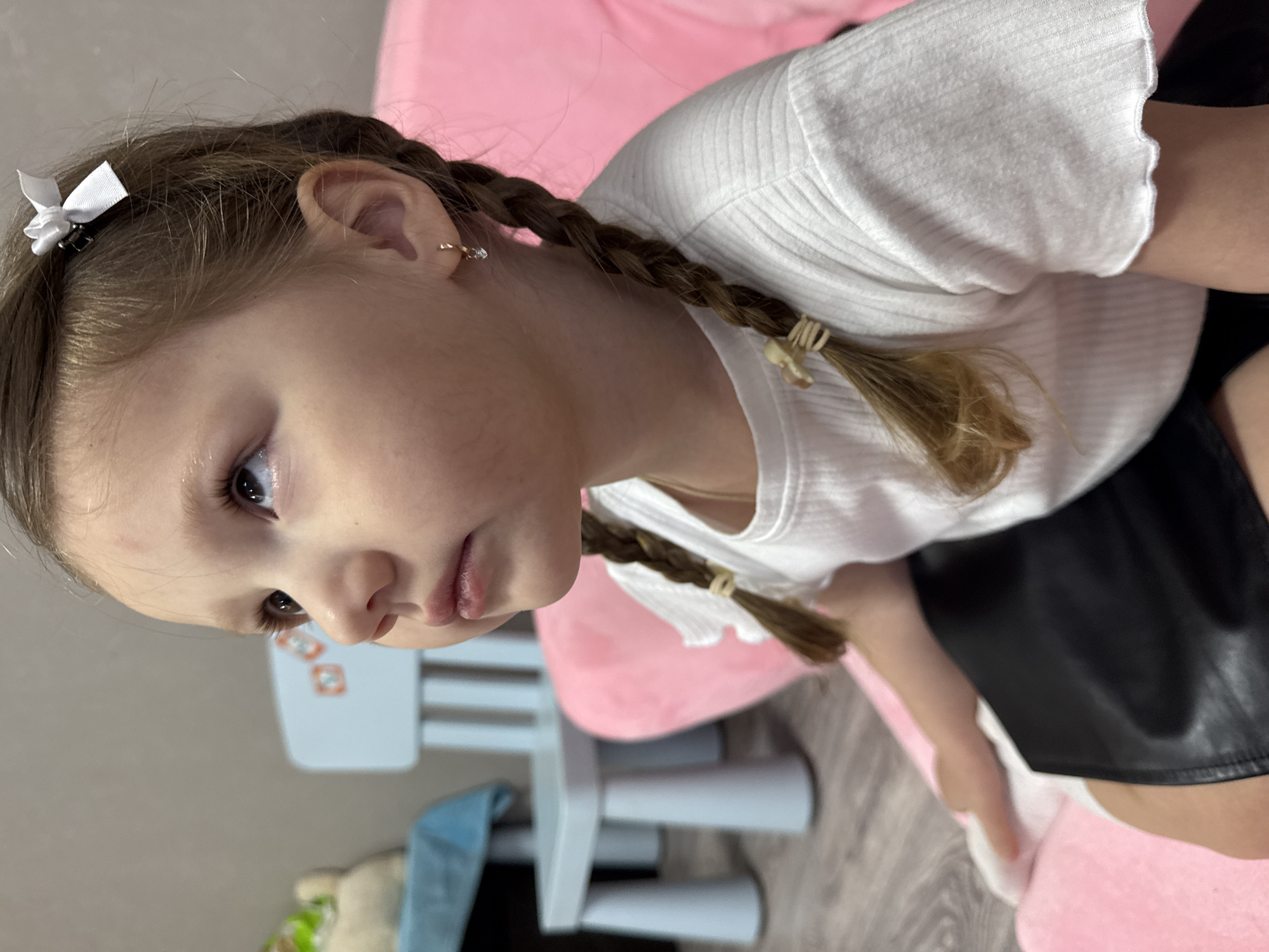 A young girl with light brown hair braided into two pigtails is captured in a close-up shot. She wears a white, short-sleeved ribbed shirt and a black skirt. Her hair is adorned with a small white bow on one side and is tied at the ends of the braids with light-colored bands. The girl has fair skin, light eyes, and a small earring in her left ear. She is seated on a pink cushion. In the background, a light blue child's chair and a plush toy partially covered by a blue cloth are visible. The location is Кипчак, Russia.