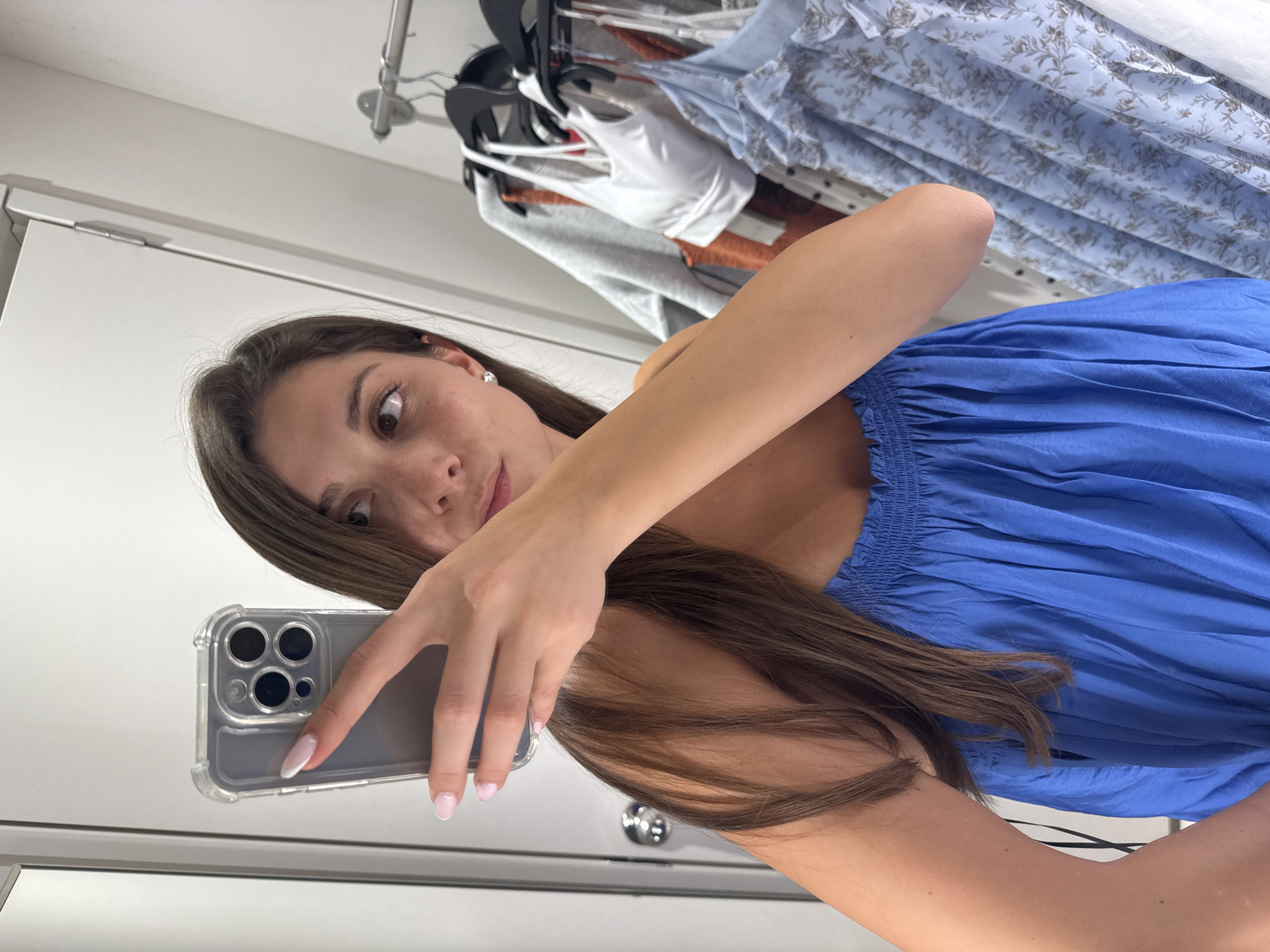 A young woman with long brown hair is taking a mirror selfie in a fitting room. She is wearing a blue strapless top with smocked detailing at the bust. Her right hand is holding up a smartphone, and her left arm is bent at the elbow, resting on her side. In the background, hangers with various clothes are visible on a rack, including a white sports bra, an orange top, and a light blue floral print dress. The fitting room appears to be in a retail store, possibly in Burgas, Bulgaria.