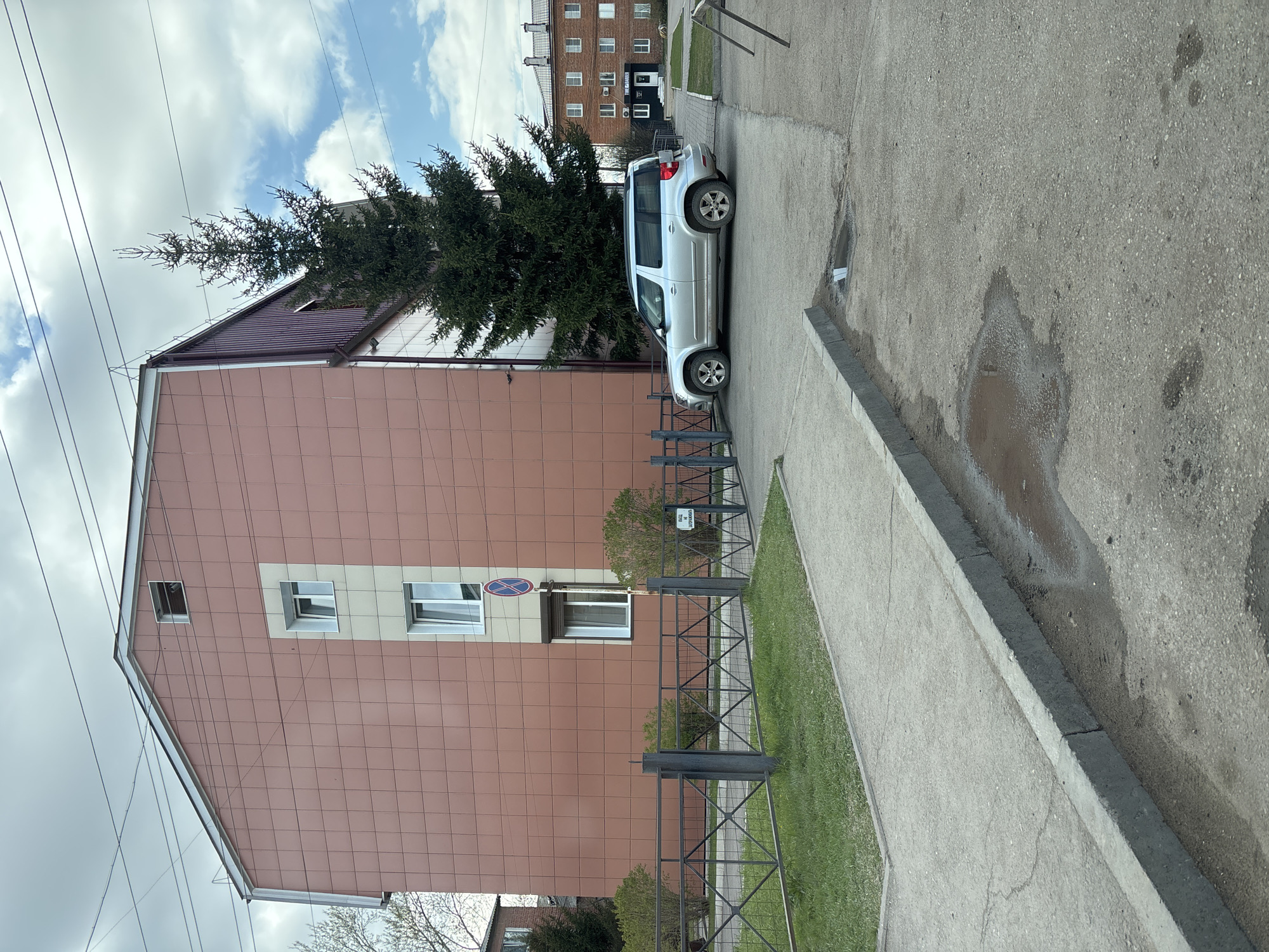 A silver SUV is parked next to a building in Ust-Ordynsky, Russia. The building is multi-story with a tiled facade and multiple windows. There is a tree next to the building and the car. A metal fence separates the building and the sidewalk. The sidewalk is concrete and there is a puddle of water on the pavement. Power lines are visible in the sky.