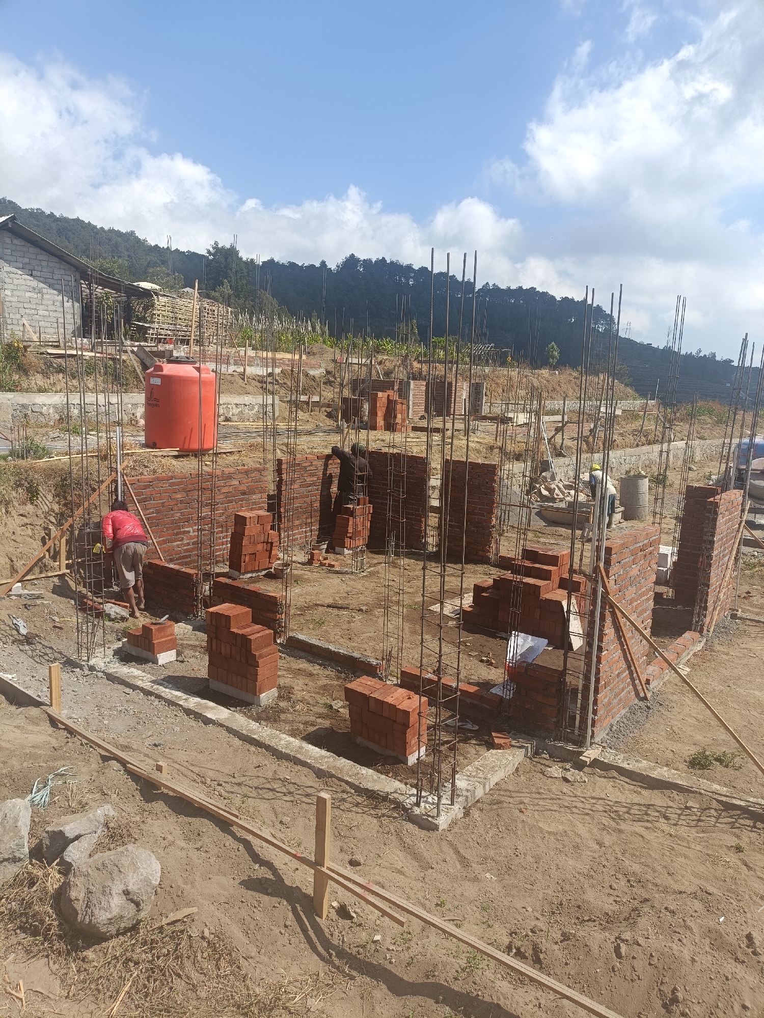 In Kopeng, Indonesia, this image depicts a construction site under a bright, cloudy sky. The foreground is dominated by loose soil and scattered rocks, with wooden stakes and planks indicating the layout of a foundation or walkway. In the middle ground, the construction of brick walls is underway. Several stacks of red bricks are visible, some neatly piled, others in the process of being laid. Reinforcement bars, essential for structural integrity, protrude from the ground and are integrated into the partially built walls.

Two construction workers are present. One, wearing a red jacket and light-colored shorts, is actively working on laying bricks at the base of a wall. Another worker, dressed in dark clothing, is positioned further back, also engaged in the construction process, appearing to be near a pile of bricks and tools.

A large, orange water tank stands prominently behind the workers, serving a practical purpose on the site. In the background, a partially constructed building with a cinder block facade is visible, alongside scaffolding and more construction materials. The scene is set against a backdrop of a densely forested hillside under a blue sky with scattered white clouds, characteristic of the Indonesian landscape. The overall impression is one of active development in a natural, hilly environment.