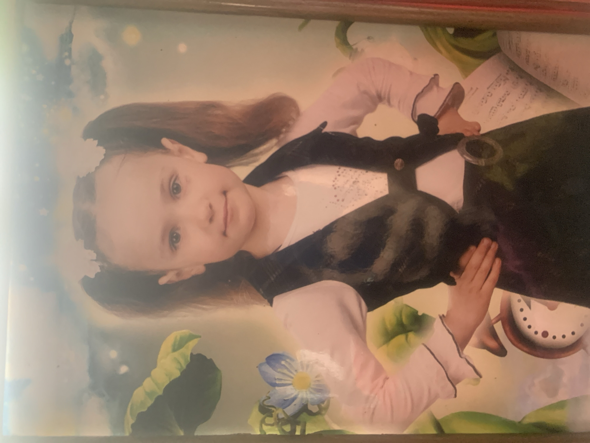 This media file is a photograph of a young girl dressed in a costume, possibly for a theatrical performance or a themed photoshoot, with a whimsical, dreamlike background. The girl, with brown hair styled in pigtails adorned with white flowers, is looking directly at the camera with a gentle smile. She is wearing a white shirt with ruffled sleeves and a dark vest over it. Her left hand is placed on her hip, and her right hand is resting on the vest.
The background features soft, diffused lighting, suggesting an indoor setting with controlled lighting. There are elements of nature, such as large green leaves and a blue flower, which contribute to the fantastical theme. To the right of the girl, a portion of an open book with text is visible, along with what appears to be a decorative clock with a brown rim and a white face with black dots.
The overall impression is one of innocence and charm, with the girl appearing confident and engaged. The image is framed by a wooden border. While the specific event or activity is not explicitly stated, the girl's attire and the artistic background suggest a playful or creative endeavor. The image itself is a photograph, likely taken in Ustyvytsia, Ukraine, given the location context. No specific time of day or weather conditions are discernible from the image.