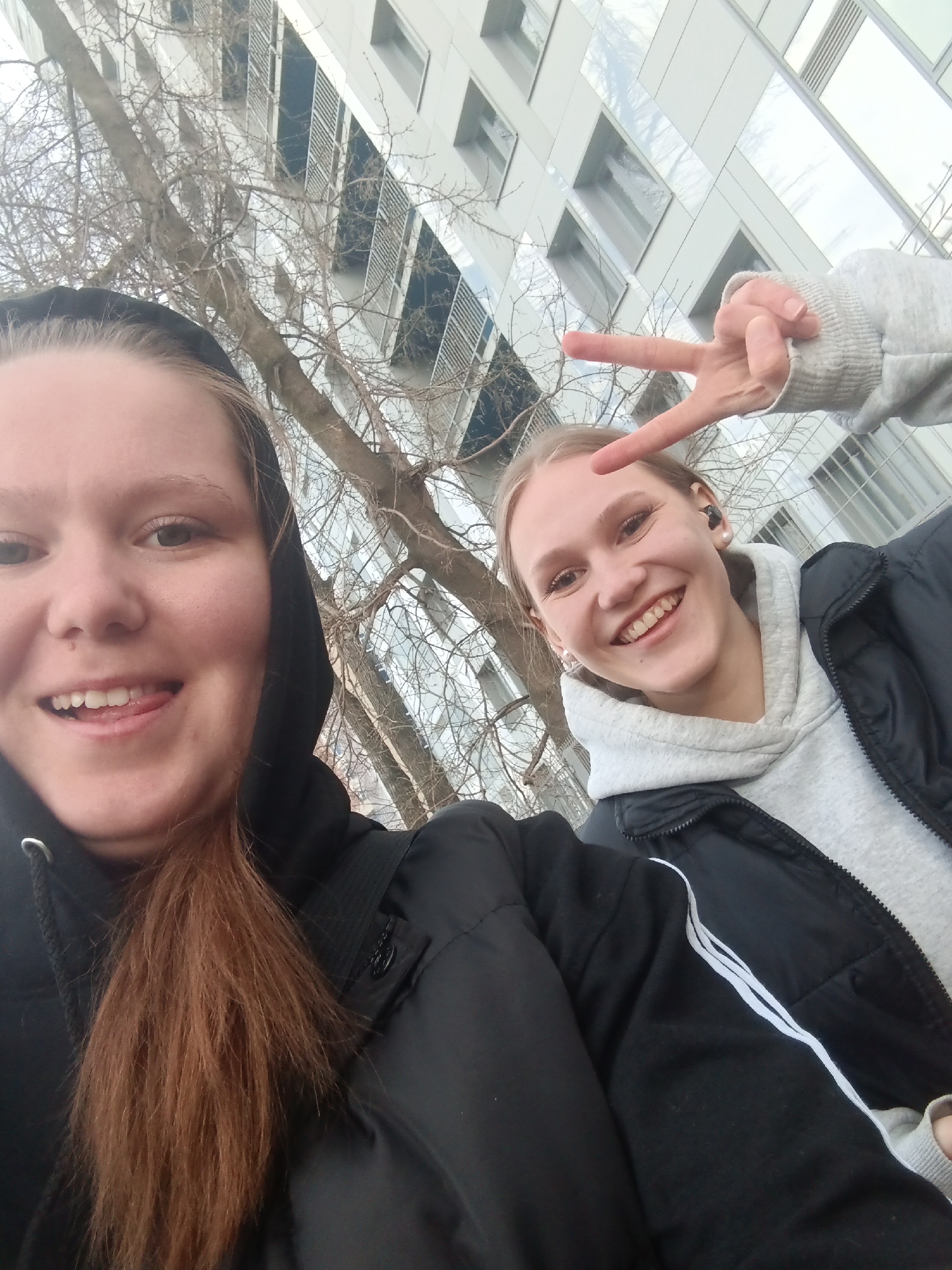 Two young women are taking a selfie outdoors. The woman on the left has red hair and is wearing a black hooded jacket. She is sticking her tongue out. The woman on the right is smiling and making a peace sign with her right hand. She is wearing a gray hoodie with a black jacket over it. The background consists of leafless trees and a modern building with a facade of mirrored or metal panels and windows. Based on the architectural style of the building and the urban setting, the location is likely in a contemporary area in Perm, Russia.