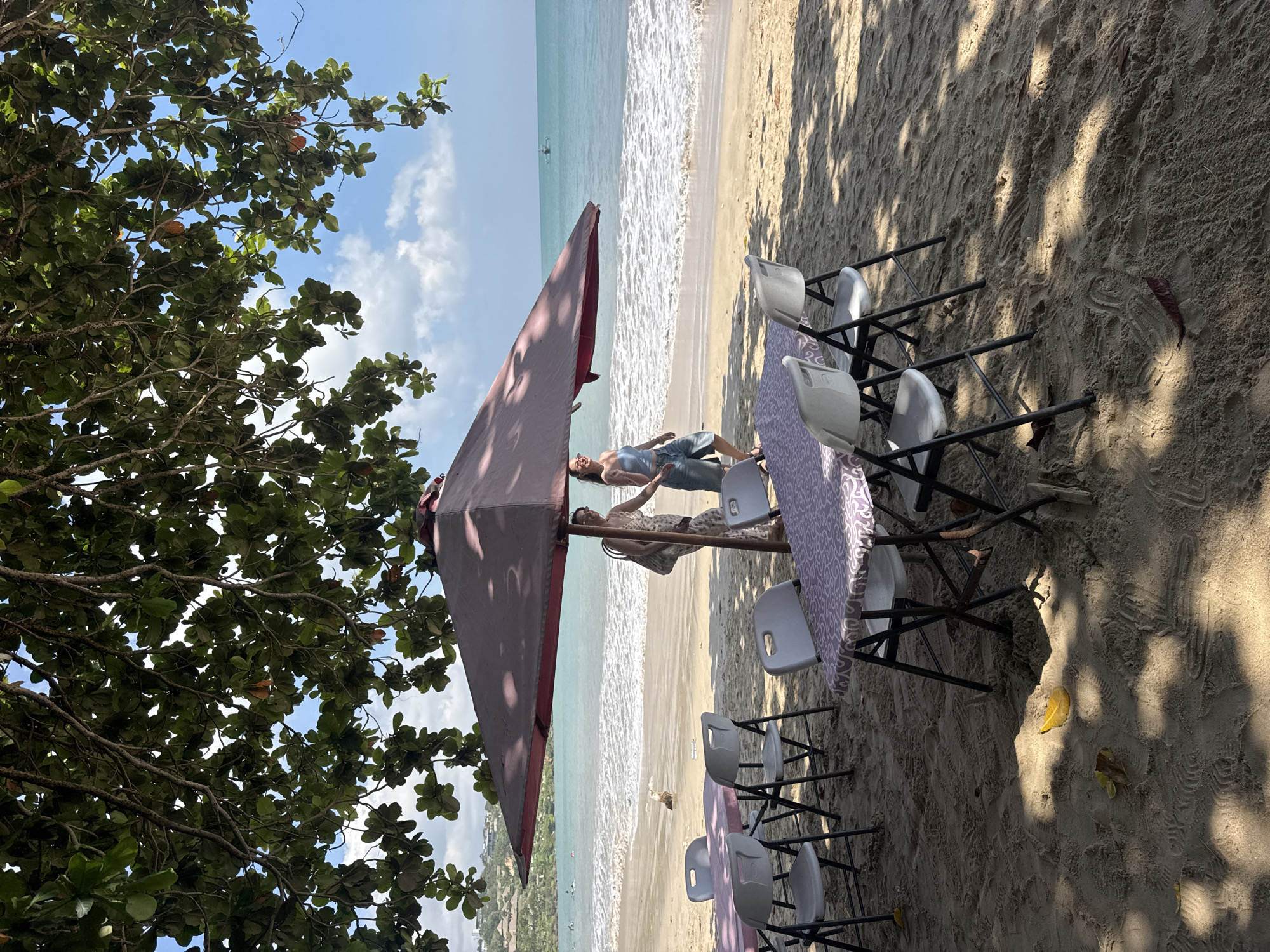 The image captures a vibrant beach scene in Kuta Selatan, Indonesia, under bright daylight. The primary setting is a sandy beach next to a tranquil blue-green ocean, with white waves gently breaking on the shore.
On the left side of the frame, a large tree with dense green foliage provides natural shade, its branches extending across the top and left of the image. Sunlight filters through the leaves, creating dappled shadows on the sand. The sky above is clear and blue with scattered white clouds.
In the mid-ground, several white folding chairs with black frames are arranged on the sand. Two tables are visible, covered with light purple patterned tablecloths, suggesting a casual outdoor dining or seating area. A large, maroon-colored beach umbrella stands prominently, casting a significant shadow and offering additional shade to a portion of the setup.
Two individuals are present near the umbrella and tables. One person, a woman with dark hair, is standing, looking towards the ocean. She is wearing a light-colored top and shorts. Another person is partially visible, seated or reclined under the umbrella, wearing a light-colored, possibly patterned outfit.
The sand shows various footprints and patterns, reflecting the activity on the beach. In the distant ocean, a small, white boat or kayak can be seen. The overall atmosphere appears relaxed and peaceful, indicative of leisure activity on a sunny day at a tropical beach. No visible text is present in the image.