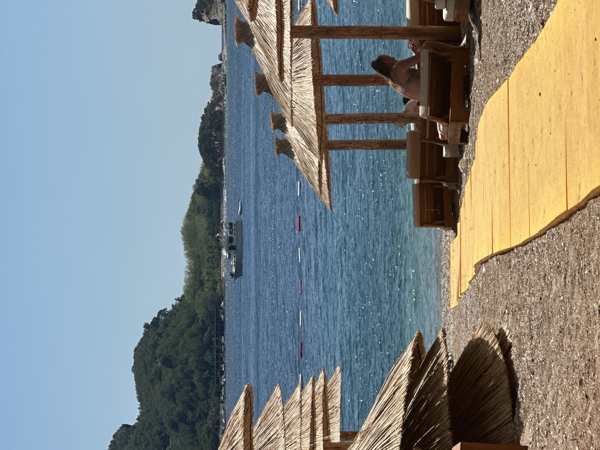 A woman wearing a bikini is sitting on a sun lounger under a thatched umbrella on a beach in Budva, Montenegro. The beach has a yellow walkway leading to the sun loungers. In the background, the blue sea stretches out to the horizon, with a boat sailing on the water. Green, tree-covered hills rise in the distance, and the sky is clear and blue. The scene appears to be a typical summer day at a coastal resort.