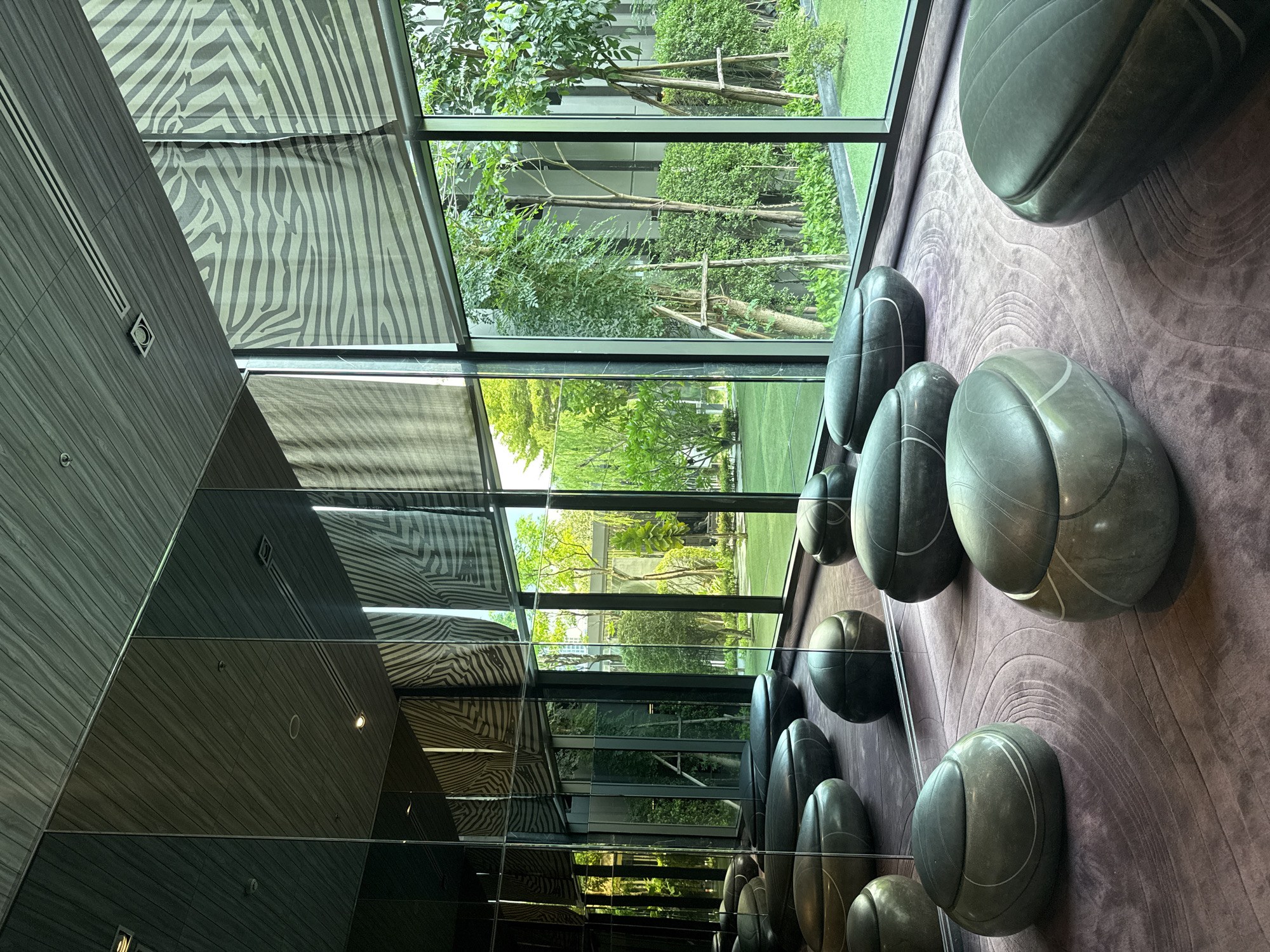 This appears to be an interior shot of a commercial or residential building in Bangkok, Thailand, with a distinctive modern design. A row of large, gray, stone-like spherical objects rests against a textured purple wall, likely part of the interior decor. The space is characterized by large windows and reflective mirrored surfaces. Through the windows, a manicured green outdoor area, possibly a garden or courtyard, is visible, featuring trees, shrubs, and a building in the distance. The windows are partially covered with zebra-striped blinds or shades. The mirrored wall reflects the zebra-striped shades and the window view, creating an illusion of depth. The overall setting is modern, clean, and upscale, suggesting a contemporary interior design.