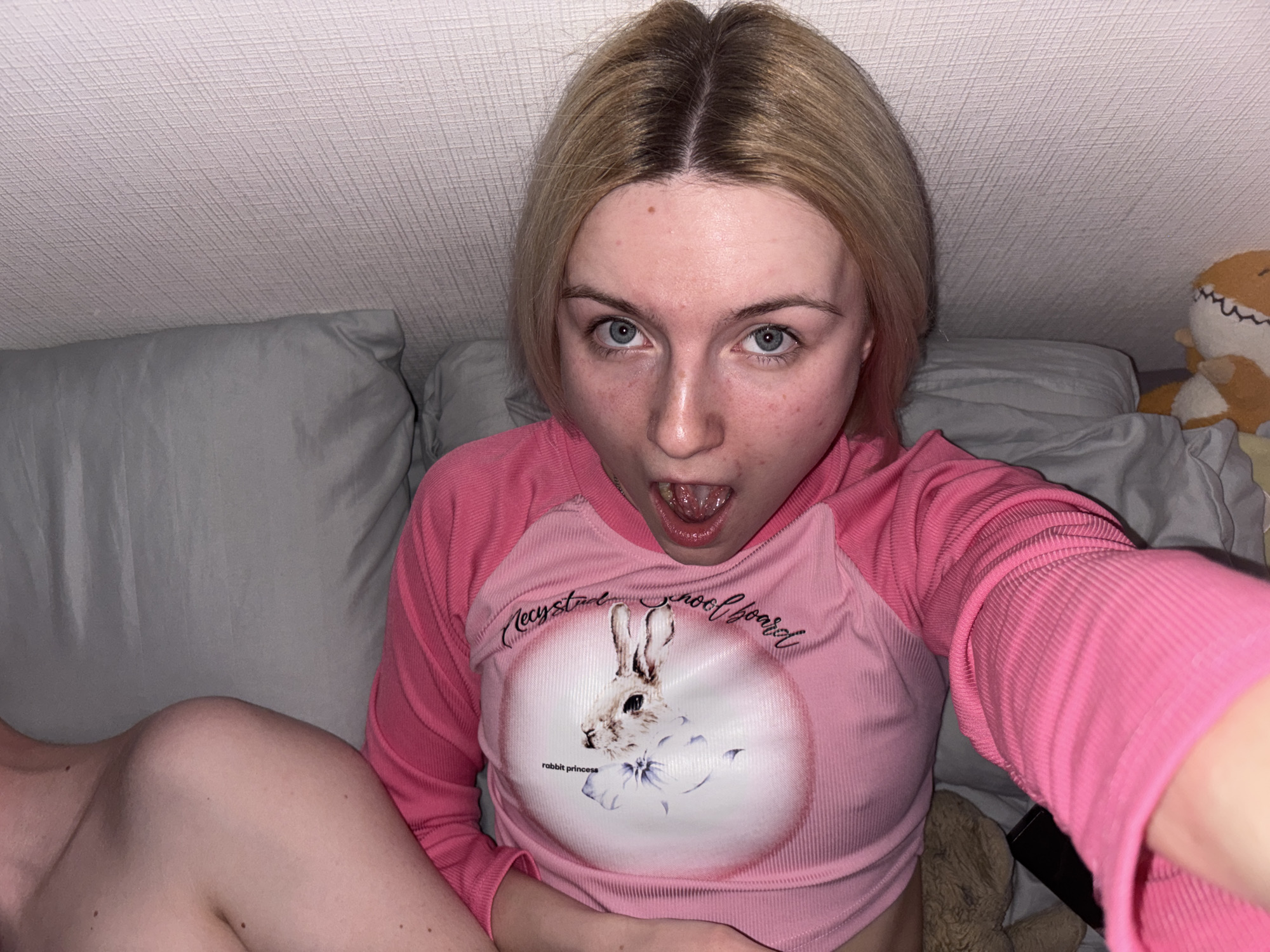 A close-up, indoor photograph features a young individual taking a selfie, displaying a surprised or playfully shocked expression with wide eyes and an open mouth. The person has fair skin, blonde hair with dark roots, and light blue-grey eyes. They are wearing a ribbed, long-sleeved pink top. The front of the shirt has a white circular graphic depicting a rabbit with a bow, above which the text "Crystal Schoolboard" is curved, and below it, "rabbit princess" is written.
The subject is reclining on grey pillows or bedding. To their left, the bare shoulder and upper arm of another person are visible, also resting on a grey pillow. The background is a light-colored, textured wall or ceiling. To the right, a large, light brown stuffed animal with stitched teeth and an eye is partially visible. Further down on the right, another fuzzy brown object and a dark rectangular item (possibly a remote) are seen. The setting appears to be a cozy indoor space, likely a bedroom. The lighting is artificial and provides no indication of the time of day. No visual cues in the image indicate the specified location of Ligolambi, Russia.