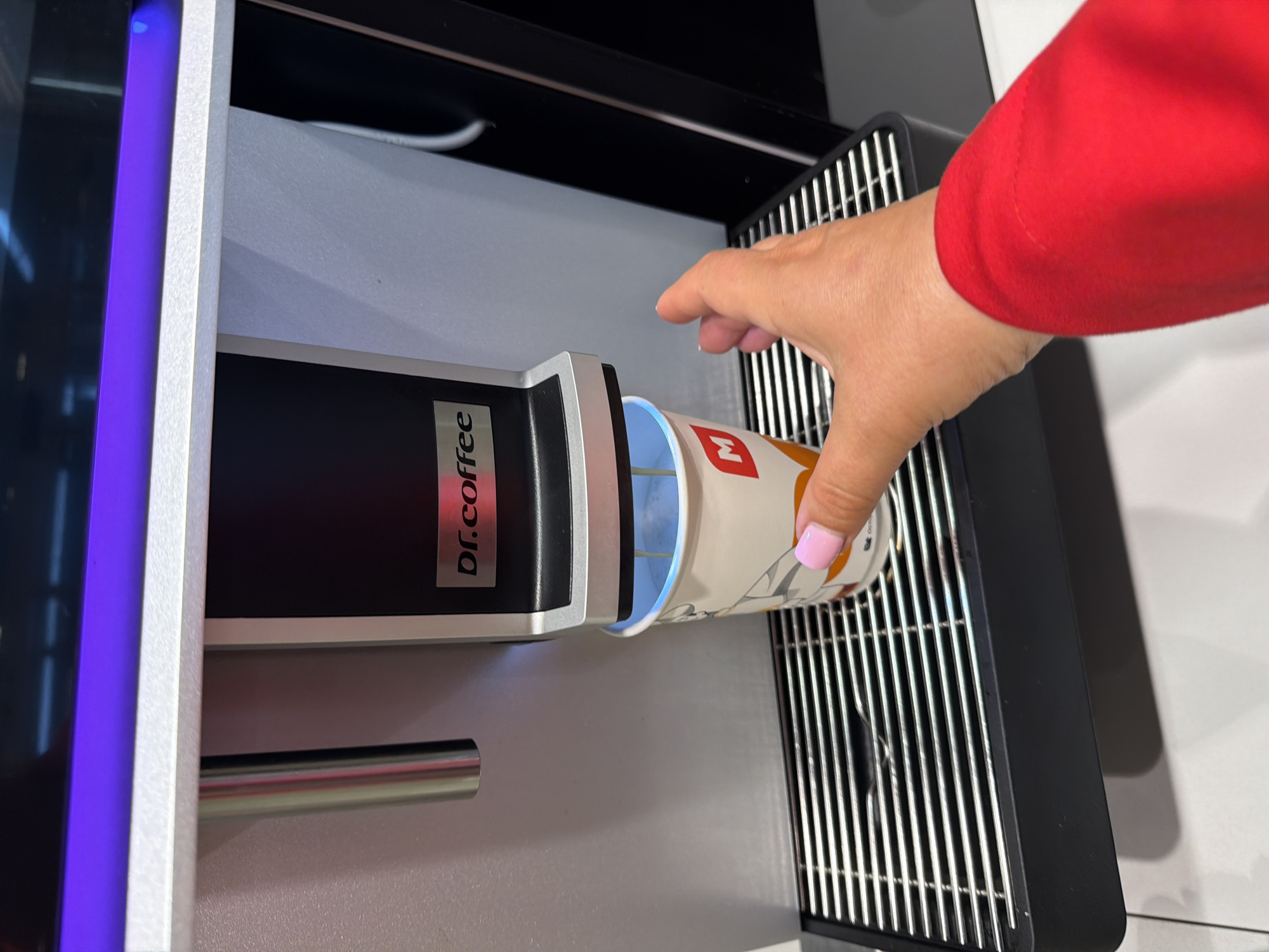 A person's hand in a red sleeve holds a paper cup with a blue interior into a "Dr. Coffee" machine, presumably dispensing coffee. The machine is silver and black with a blue light on the left side. The scene appears to be inside a building with modern appliances. There is no indication of the location in the city of Leningradskaya, Russia.