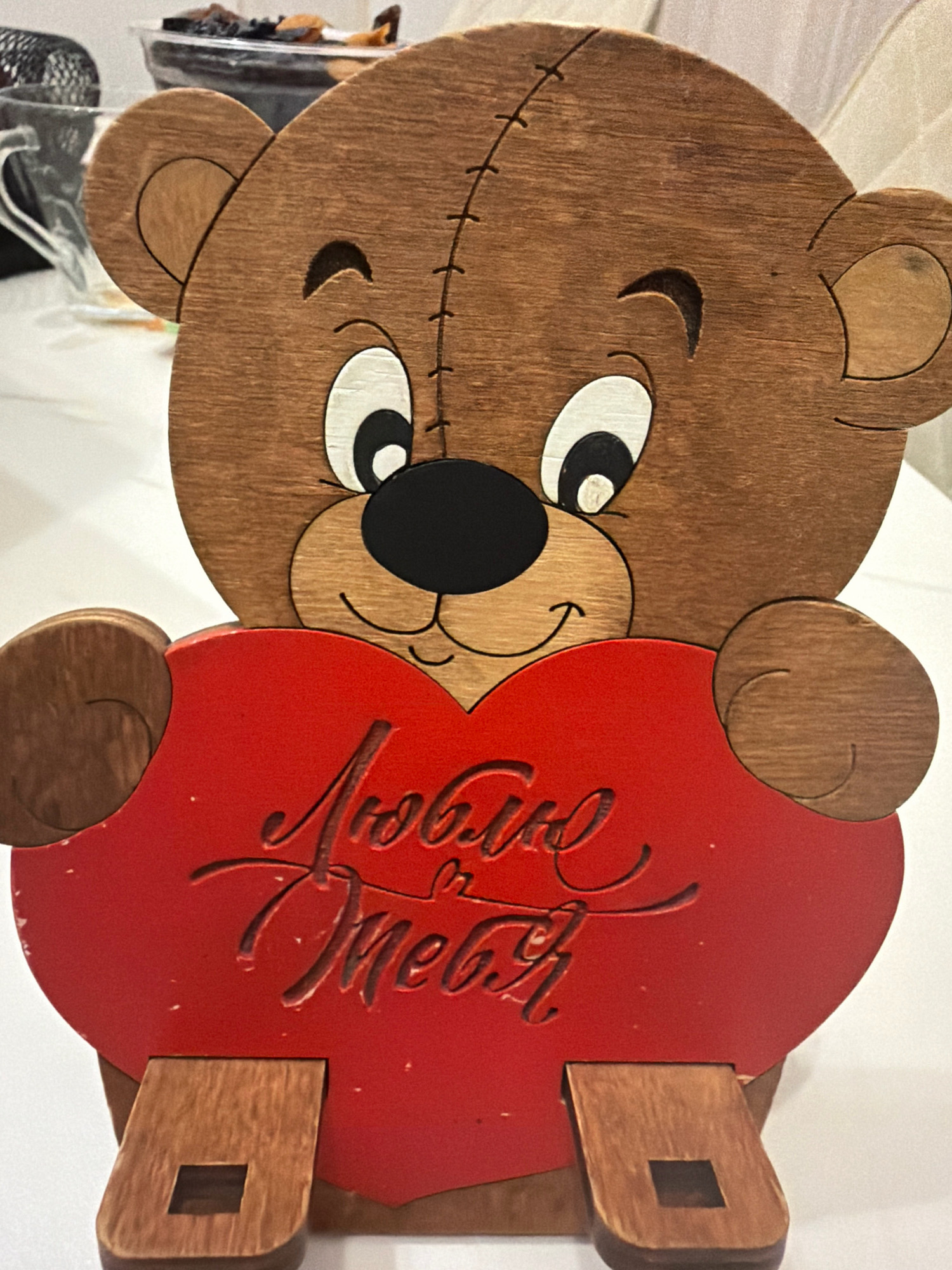 A wooden, carved teddy bear figurine with a red heart held in its paws is depicted against a white surface. The bear has stitched details on its head, large round eyes with white and black pupils, a black nose, and a smiling mouth. The red heart has cursive writing on it that appears to be in Russian. The bear is standing on two wooden legs. The background is out of focus but suggests an indoor setting with some shelves containing items. The location in Vniissok, Russia is not visible in the image.