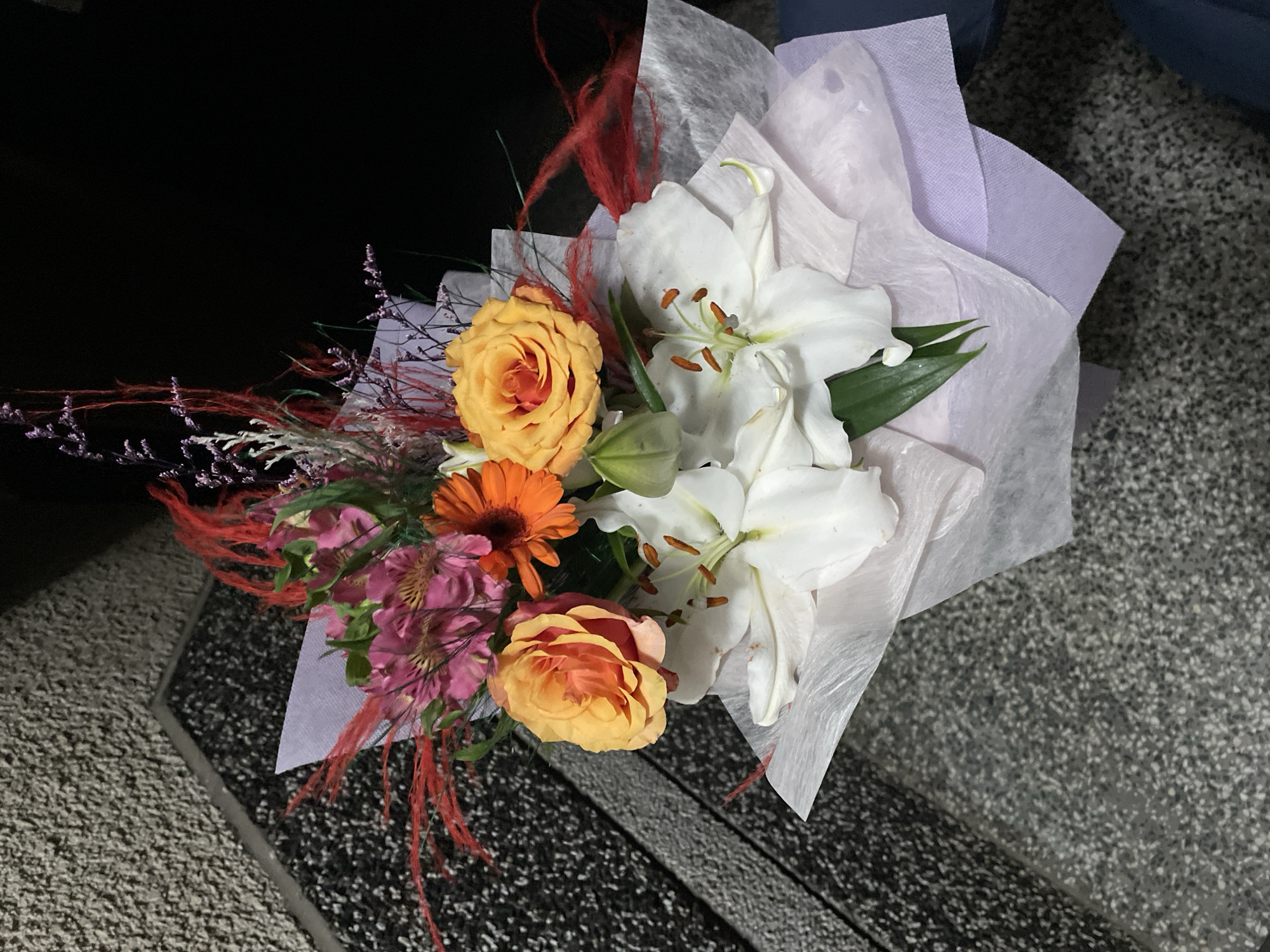 A bouquet of flowers rests on a speckled gray surface. The bouquet is composed of white lilies, peach-colored roses, a single orange gerbera daisy, and smaller pink alstroemeria. Red decorative strands extend outward from the bouquet. The flowers are wrapped in layers of white and lavender paper. The location in the city of Zlatograd, Bulgaria, is unknown.