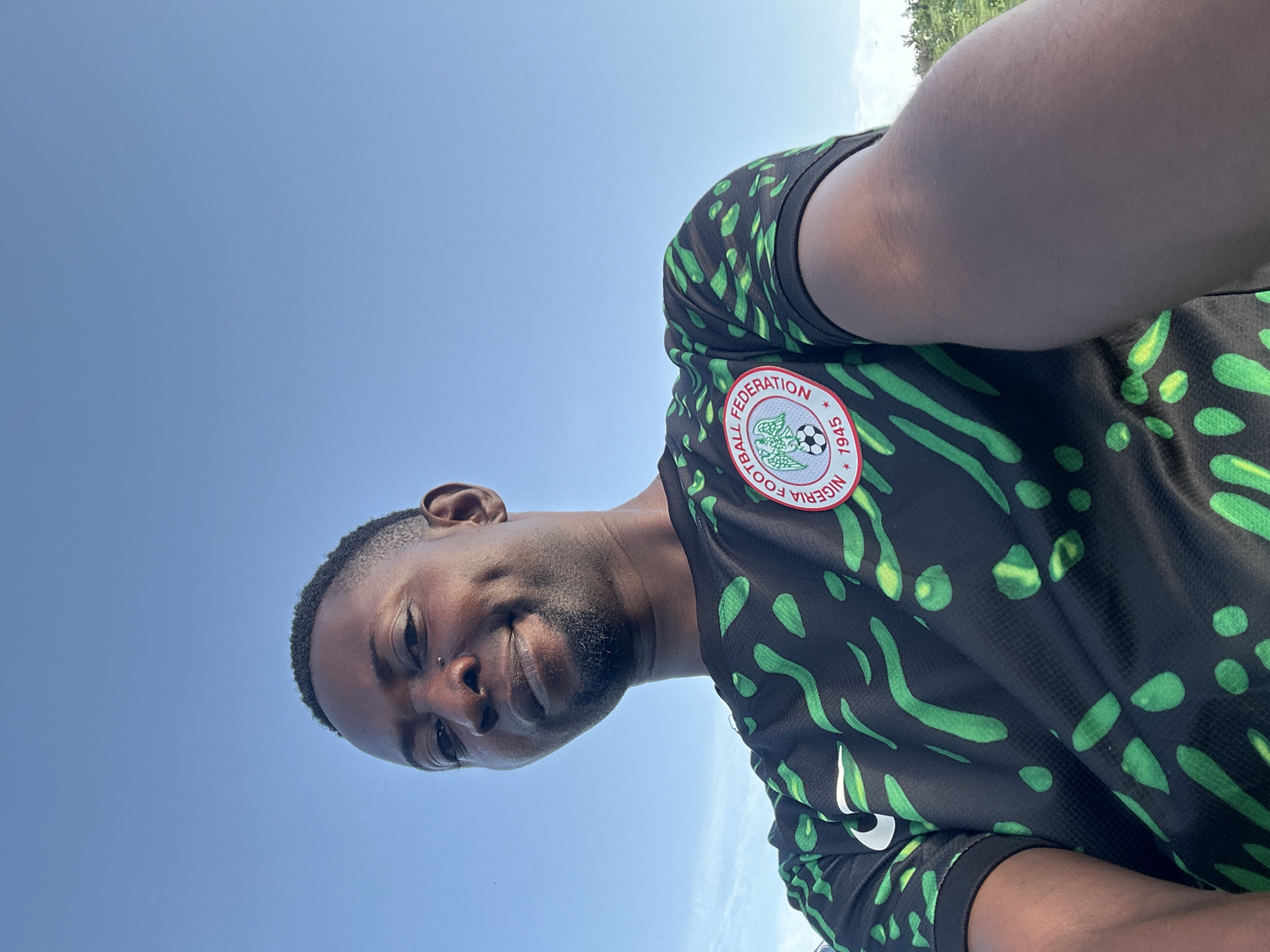 The media file is a selfie of a man wearing a black and green patterned shirt with a circular logo on the chest. The logo has text around it, and a soccer ball is visible inside. The man has dark skin, short hair, and appears to be smiling faintly. He has a small piercing on his nose. The background is a bright blue sky, suggesting it is daytime. The image is oriented sideways. The setting appears to be outdoors, possibly in Gidan Waziri, Nigeria, given the location context. The overall mood seems casual and relaxed.