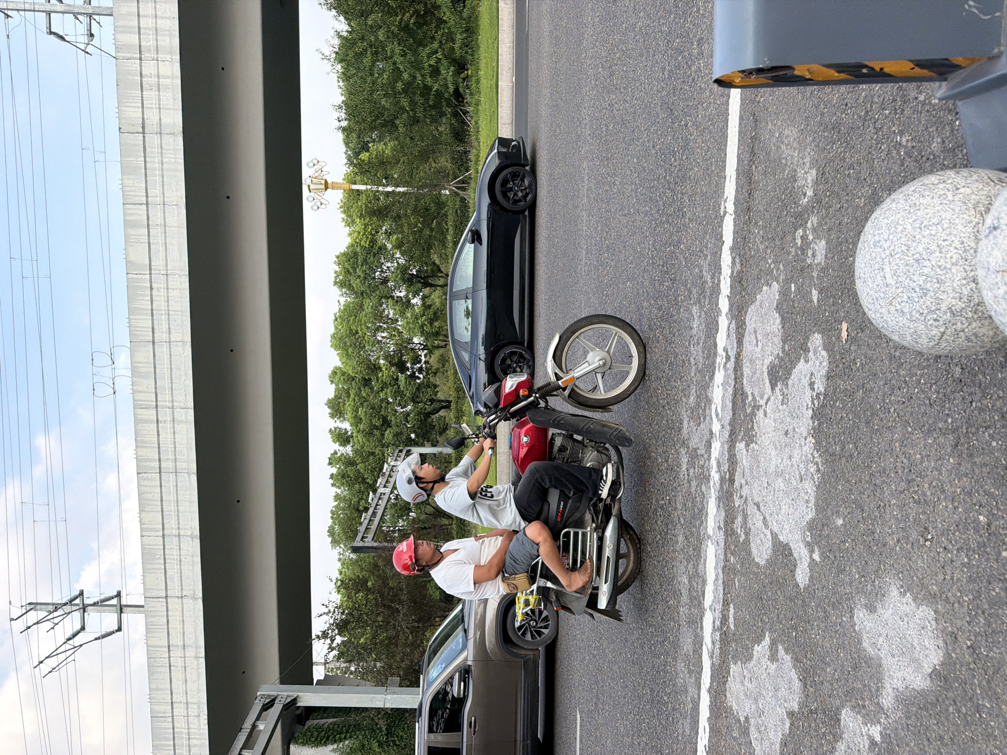 Two individuals are riding on a red motorcycle on a paved road, which appears to be a highway or main thoroughfare in Jiangjin, China. The person in the front is operating the motorcycle, while the person in the back is a passenger. Both are wearing helmets. Behind them, a dark-colored sedan is driving in the same direction. Further in the background, a beige SUV is parked on the side of the road. Overhead, an elevated concrete structure, likely an overpass or bridge, supports power lines. The environment is characterized by green trees lining the side of the road and a cloudy sky. The road surface shows some wear and tear with patches of lighter gray material.
