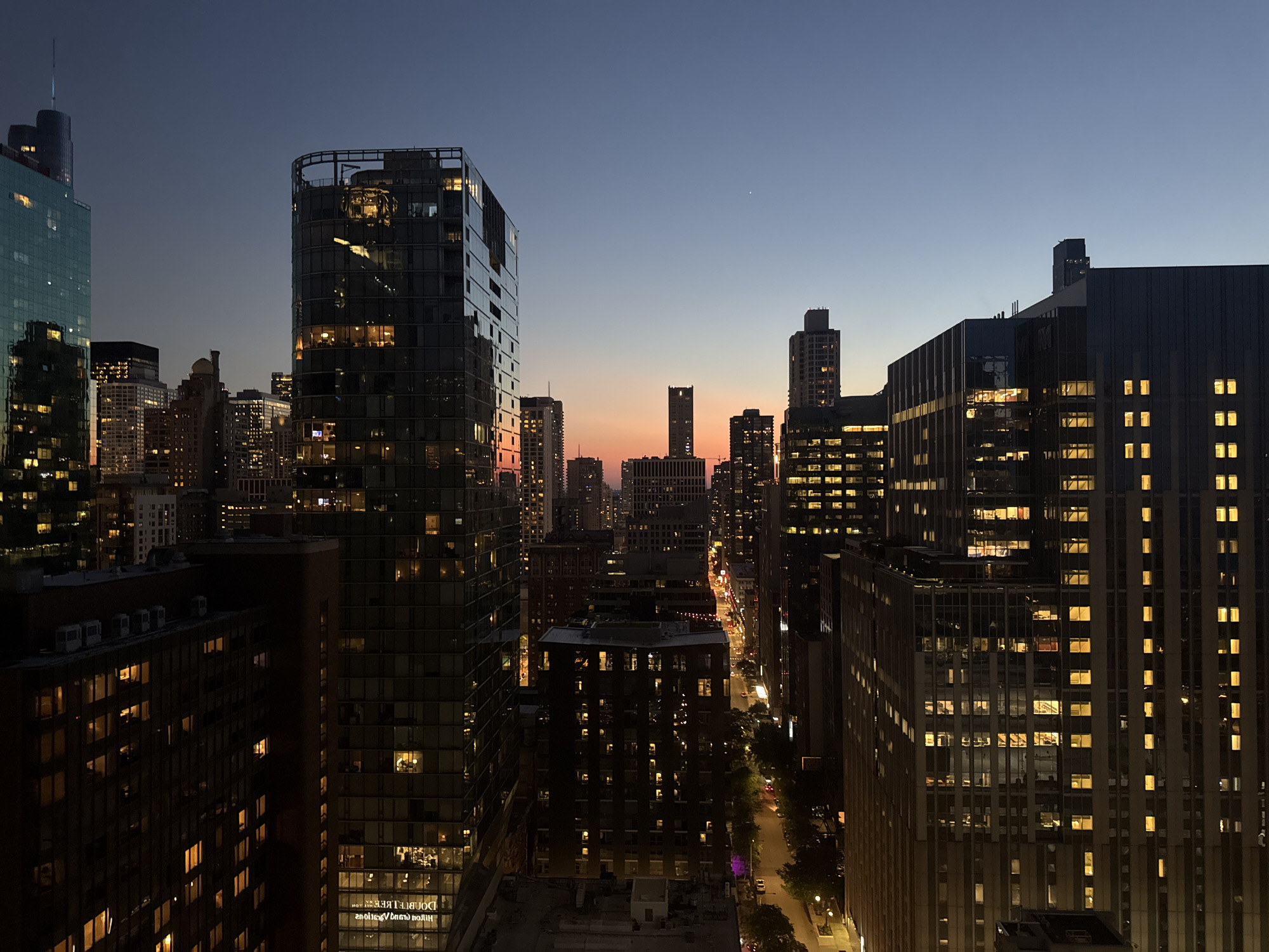 This is a high-angle, wide-shot photograph of the Chicago skyline at dusk, taken from a building looking towards the east. The sky transitions from a deep navy blue at the top to a soft orange and pink hue near the horizon, indicating the sun has recently set. The city is illuminated by the warm glow of lights from numerous high-rise buildings.
In the foreground, to the left, is a modern, dark-colored glass skyscraper with a distinctive rounded top structure. To its right, a taller, rectangular glass skyscraper with a dark frame dominates the mid-ground. Numerous windows on these buildings are lit, revealing interior spaces. Further into the cityscape, other skyscrapers of varying heights and architectural styles are visible, many with illuminated windows. A street is visible in the distance, with car headlights and streetlights creating streaks of light. Trees line the street, and some smaller buildings with lit windows are also present. The overall impression is of a bustling urban environment transitioning into night. There are no visible people or discernible actions.
