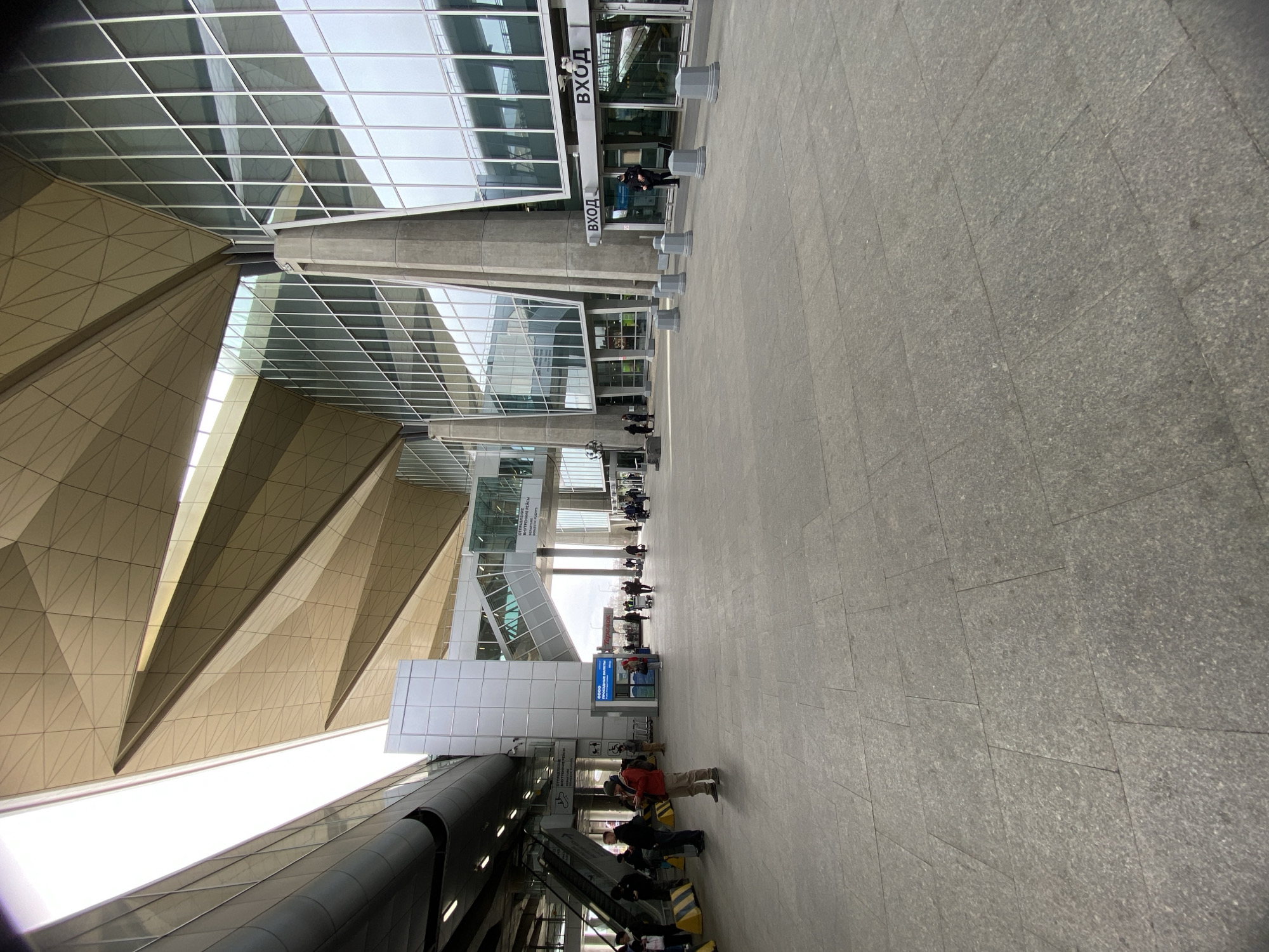 This image captures the exterior of a modern airport terminal in Saint Petersburg, Russia, taken from a slightly low angle and showing a wide perspective of the entrance area. The architecture is distinctive, featuring a large glass curtain wall structure combined with geometrically faceted, light-colored panels that form an imposing canopy or roof section. The ground is paved with large, uniform grey rectangular tiles.
Numerous people are visible throughout the scene, engaged in various activities related to travel. Some individuals are walking across the tiled expanse, while others are congregating near the building's entrances or ascending an escalator located on the left side of the frame. Many are carrying luggage, reinforcing the setting as an airport. A series of grey cylindrical bollards or barriers lines the approach to the building.
The scene appears to be set during daytime, under overcast conditions or diffused lighting, as there are no harsh shadows. Several signs are affixed to the building, with prominent text in Russian. "ВХОД" (Entrance) is clearly visible above multiple glass doorways. Other partial texts include "ОТПРАВЛЕНИЕ РЕЙСЫ" (Departures Flights), "ВНУТРЕННИЕ РЕЙСЫ" (Domestic Flights), and "ДОМАШНИЕ РЕЙСЫ" (Domestic Flights), indicating specific areas or services within the terminal. A blue kiosk bearing the "ТЕЛЕ2" (Tele2) mobile operator logo can also be seen. The overall impression is that of a bustling, contemporary transit hub.