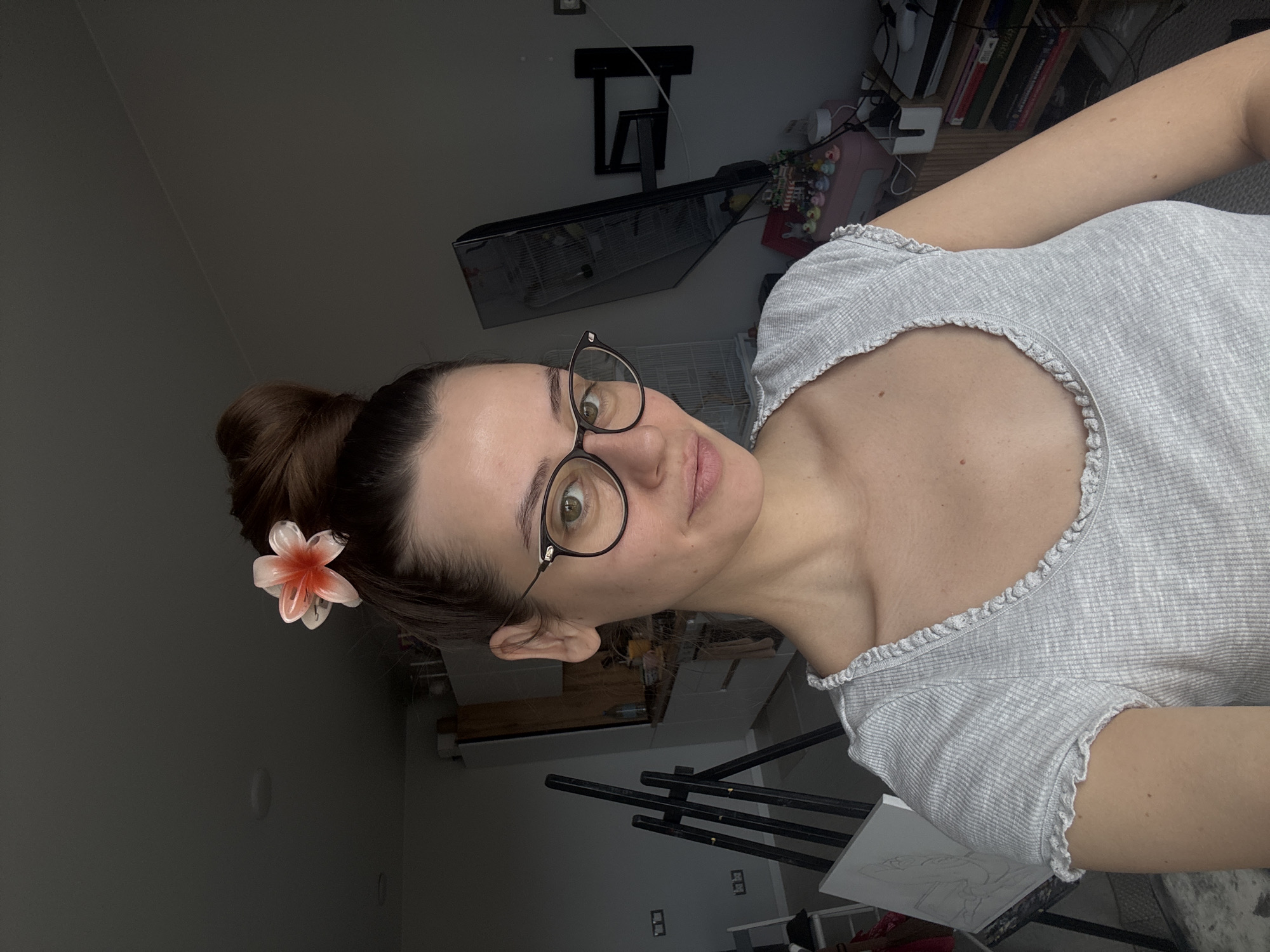 A woman with brown hair pulled back in a bun with a white and pink flower hair clip poses for a selfie. She is wearing black-rimmed glasses and a light gray ribbed shirt with a ruffled neckline.
Behind her is a room with an easel holding a canvas, a birdcage hanging from the wall, shelves of books and various items, and a partially visible kitchen area. The location in the city of Krasnoyarsk, Russia, cannot be determined from the image.