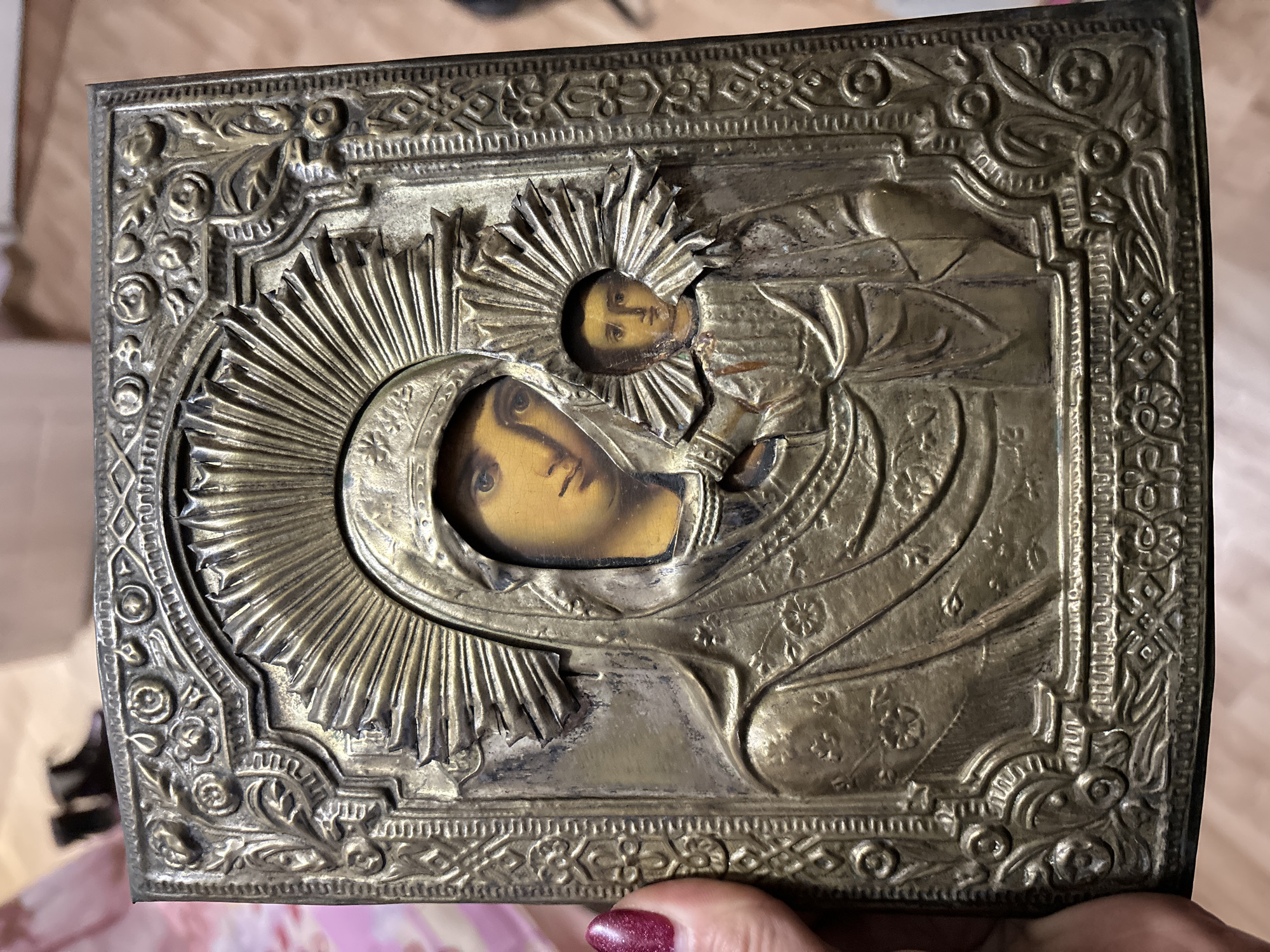 A tarnished, rectangular metal basma (oklad) covers an Orthodox icon depicting the Virgin Mary and Christ Child, held vertically by a human hand. The basma, likely of brass or bronze, features embossed patterns including floral motifs, geometric borders, and radiating halos around the painted faces and hands of Mary and the Child. The visible skin tones in the painted areas are yellowish. The metal shows extensive tarnish, discoloration, and minor deformation, particularly around the Christ Child's halo. A left human thumb, adorned with dark glittery red nail polish, is visible gripping the icon's left edge. The background is blurred, indicating an indoor setting with indistinct wooden surfaces and other dark objects. This scene is located in Chaikovsky, Russia.
