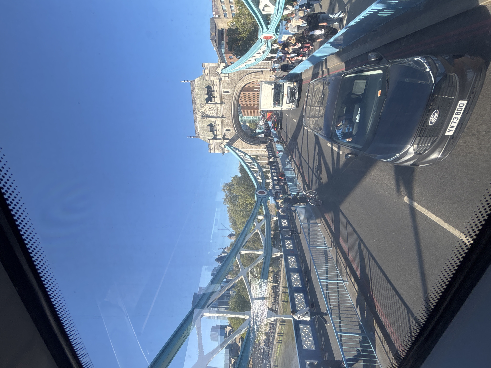 The image captures a vibrant, sunny daytime scene on Tower Bridge in London, United Kingdom, viewed from the interior of a vehicle, likely a bus, looking slightly downwards through its windshield. The perspective is tilted, showing the road and bridge structures vertically.
In the foreground, part of a vehicle's windshield with a dotted frit band is visible. The bright blue sky fills a significant portion of the upper left, indicating clear weather.
The main subject is Tower Bridge, with its distinctive blue and white structural elements and grand stone towers. On the roadway, traffic is moving. A prominent black Ford SUV or van is seen in the midground, with its front grille, "Ford" emblem, and license plate "YV73 BDO" clearly visible. Behind it, a white truck and other vehicles are present. A cyclist can also be seen on the road. Pedestrians are walking on the pedestrian walkways of the bridge, which run parallel to the vehicular traffic.
Further into the background, the ornate stone architecture of the bridge's towers stands out against the clear sky, with buildings visible beyond the bridge, suggesting an urban environment. Glimpses of water, likely the River Thames, can be seen beneath the bridge, reflecting the sunlight. Lush green trees are visible on the riverbanks.
The scene depicts a busy day with active vehicle and pedestrian traffic on an iconic London landmark. Strong shadows are cast across the road and bridge structures, indicating bright sunshine. The overall mood is bustling and bright.