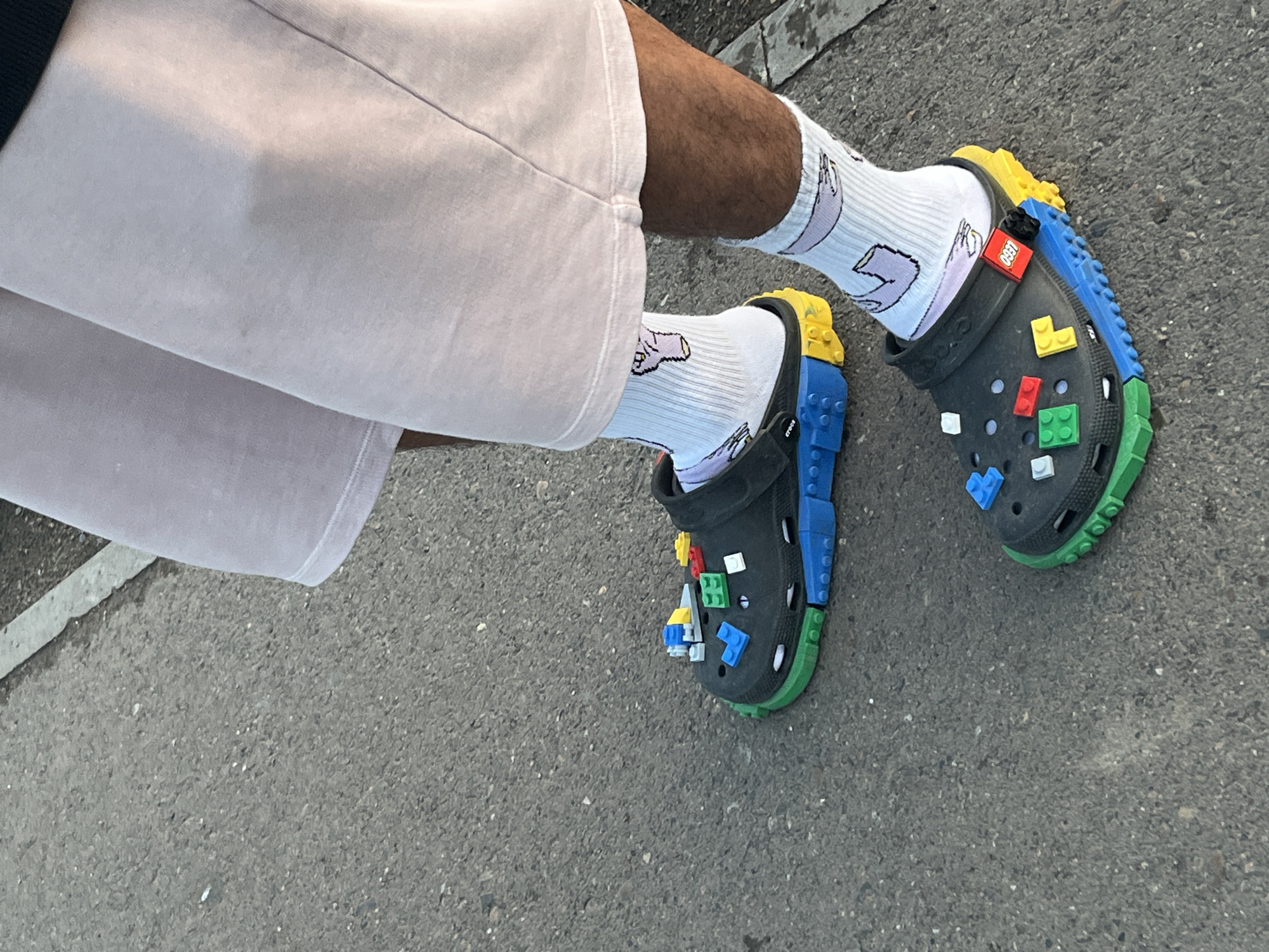 The image shows a person's legs from the knees down, standing on a gray asphalt surface with some white gravel. The person is wearing light pink shorts and white socks with a cartoon drawing of a hand giving the middle finger. On their feet, they are wearing black Crocs. The Crocs are heavily customized with what appear to be LEGO bricks. The sole of the Crocs has been augmented with colorful layers of LEGO-like pieces in blue, green, and yellow. Several smaller LEGO bricks in red, blue, green, yellow, and white are attached to the top of the shoes. One shoe has a red LEGO brick with "LEGO" written on it. The setting appears to be an outdoor urban environment, possibly a sidewalk or street. There is no specific indication of the location being in Krasnoyarsk, Russia.