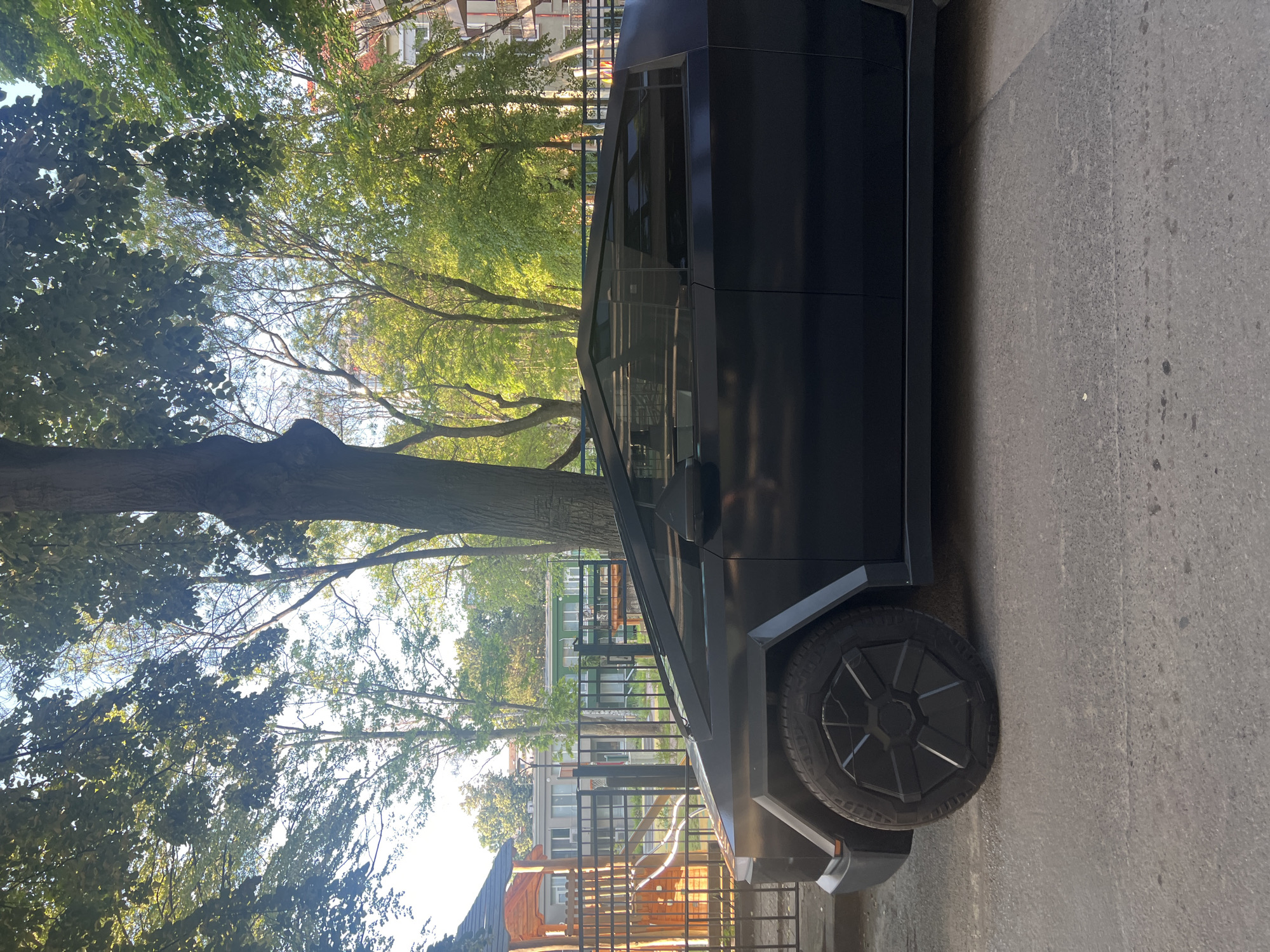 A matte black Tesla Cybertruck is parked on a paved street in front of a black metal fence. Behind the fence, a children's playground with a wooden play structure is visible, as well as several multi-story buildings and trees. The sky is visible through the leaves of the trees. The scene is captured in daylight, with sunlight filtering through the trees, creating dappled light on the ground and the vehicle. The specific location is stated to be Sofia, Bulgaria. No people are visible in the image.
