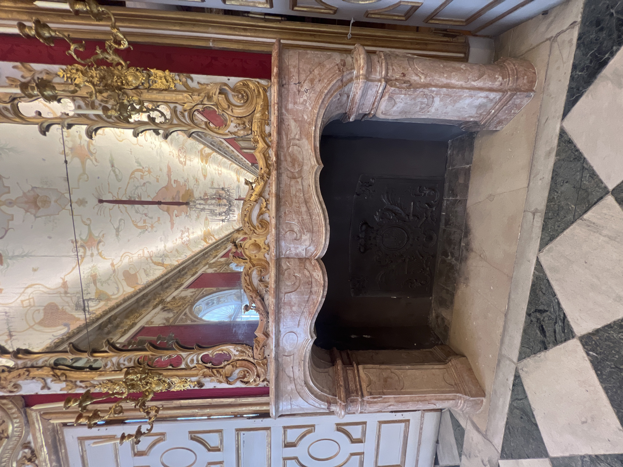 The image shows a grand fireplace in a richly decorated room, likely part of a palace or stately home. The fireplace is made of pinkish marble with ornate carvings. Above it is a large, gilded mirror framed with intricate golden scrolls, reflecting a high ceiling adorned with elaborate frescoes and a chandelier. The walls feature red drapes and decorative panels, and a gilded candelabra is visible on the left. The floor is tiled in a diamond pattern of black and white squares, with some areas of green marble. This scene is located in the New Schleißheim Palace in Oberschleißheim, Germany.