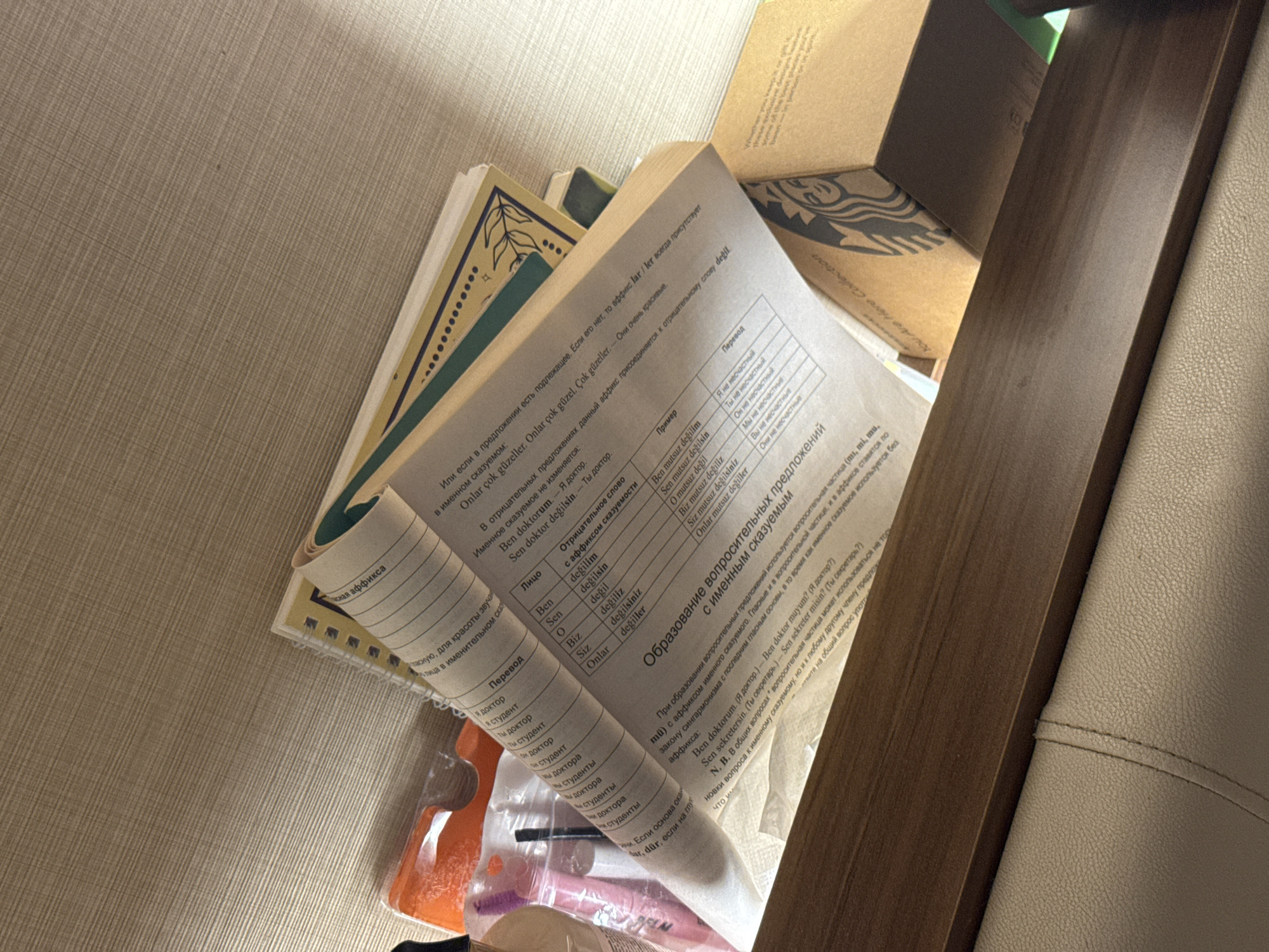 The image shows a stack of books and other items on a shelf, with a partial view of a person's hands. The books appear to be educational materials, possibly language learning books, with text in Russian and Turkish. One of the books has a section on Turkish grammar, discussing suffixes and sentence structure. There are also some makeup items and a Starbucks cup visible in the scene. The lighting suggests an indoor setting, possibly a home or an office. The overall impression is one of a cluttered but functional workspace. It is not possible to determine the location in the city of Aksu, Turkey.