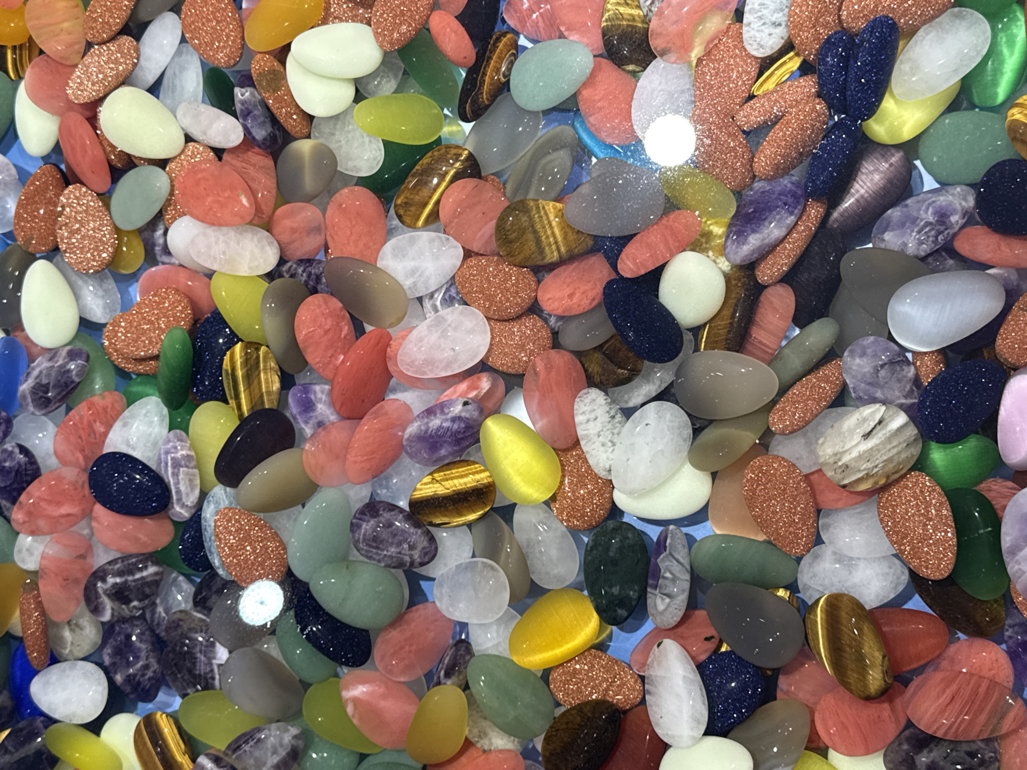 The image displays a close-up, overhead view of a collection of polished, tumbled stones. The stones vary in shape, size, and color, with hues of pink, orange, yellow, green, blue, purple, white, and brown. Some stones have a glittery, metallic sheen, while others are translucent or opaque. They are arranged in a dense, overlapping pattern against a light blue background, suggesting they are in a container or spread out on a surface. There are no people, actions, or interactions visible in the image. The location of Yuzhong District, China is not discernable from the visual information presented.